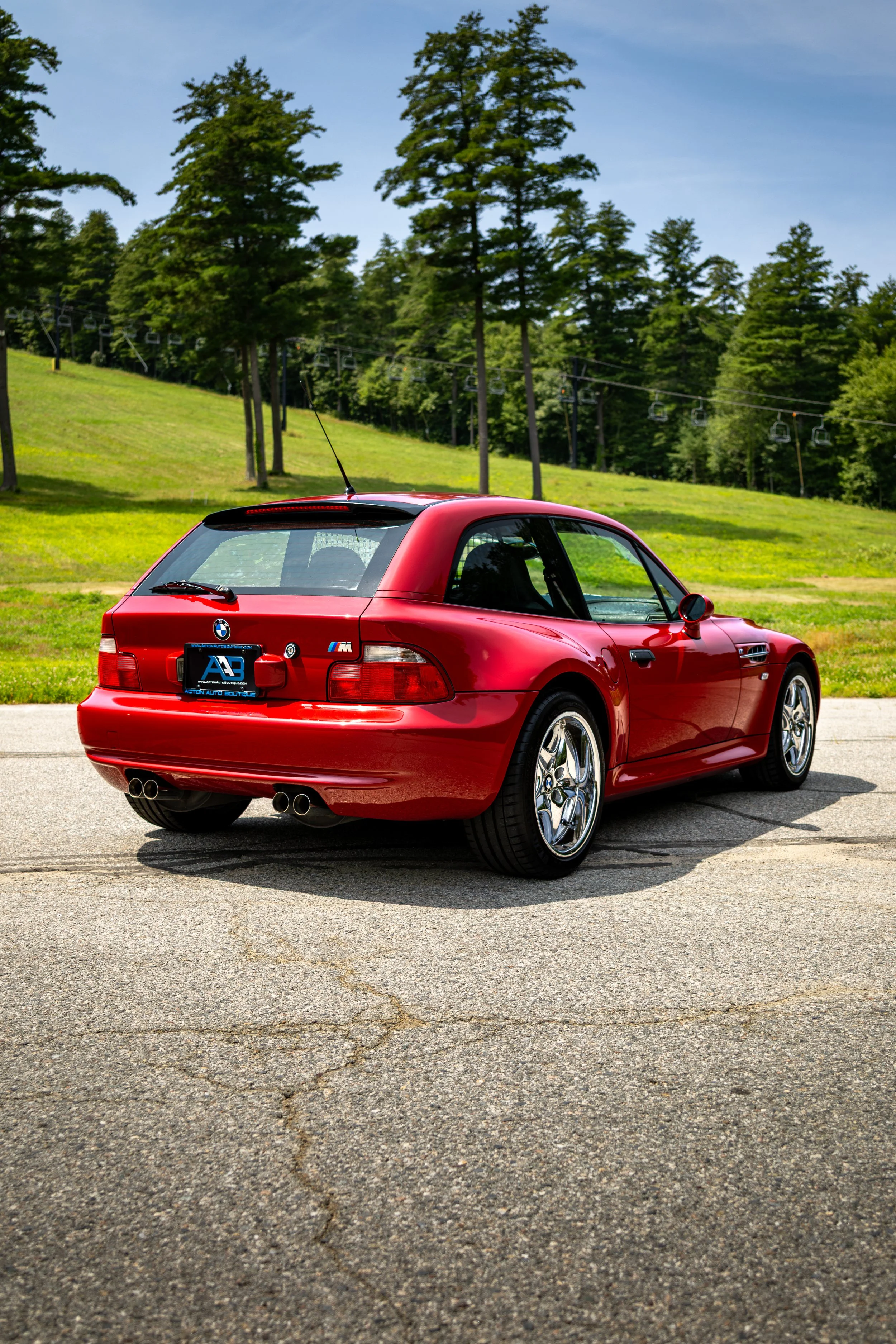 Red BMW M coupe parked on a paved surface with green grass, trees, and a blue sky in the background.