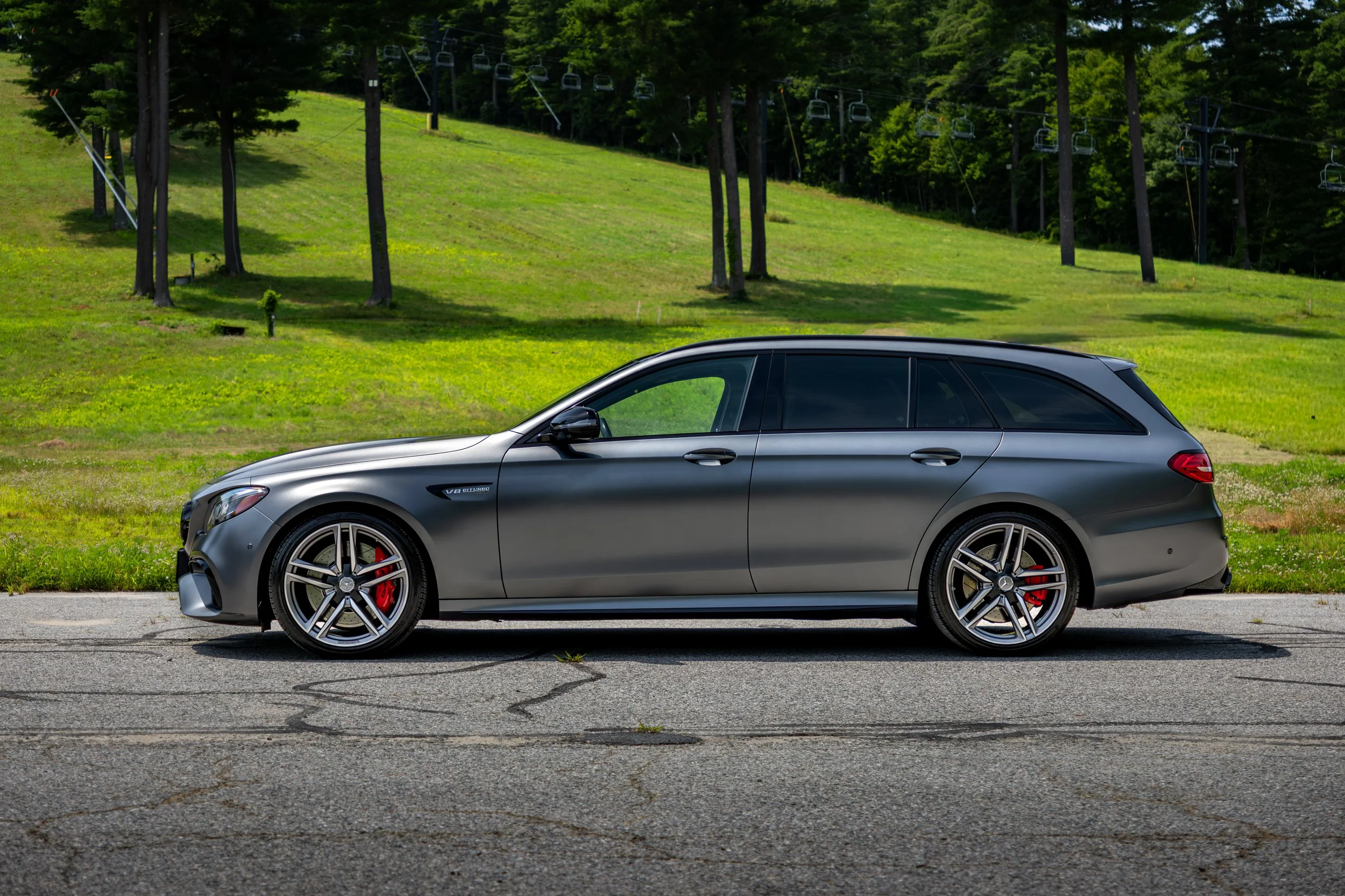 A sleek flat gray 2018 Mercedes E63s AMG wagon parked on a paved road in front of a grassy hill with trees and ski lift chairs.