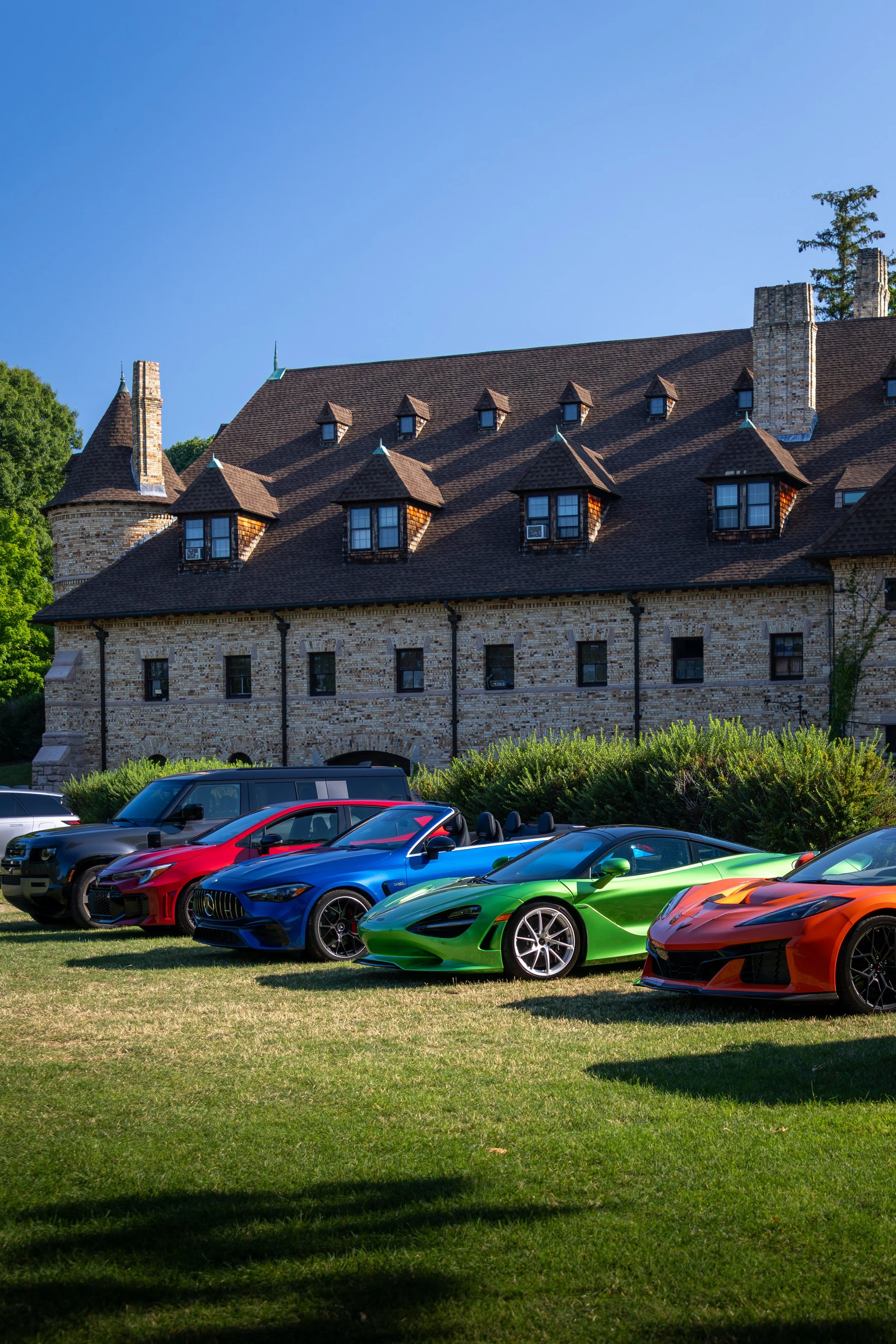 Line of colorful sports cars parked on a grassy area in front of a large brick building with turreted towers and dormer windows (the Larz Anderson Automotive Museum in Brookline, MA), with trees and a clear blue sky in the background.