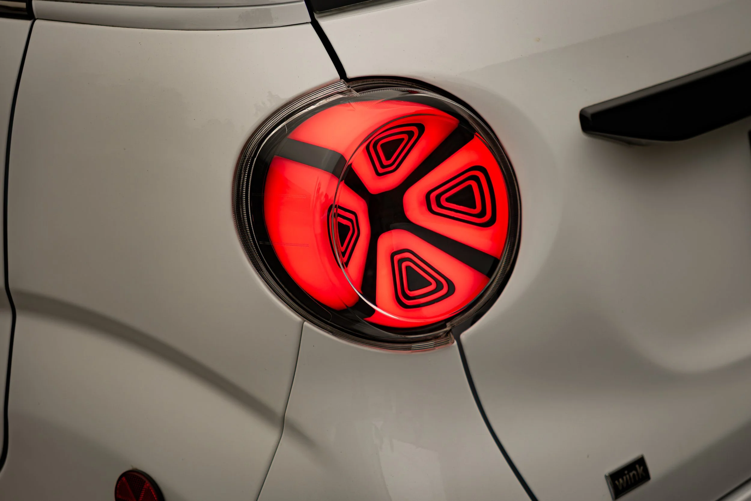 Close-up of a Wink's rear light with an illuminated red and black circular rear taillight, reflecting the car's body, with a small black sticker labeled 'wink' visible in the lower right corner.