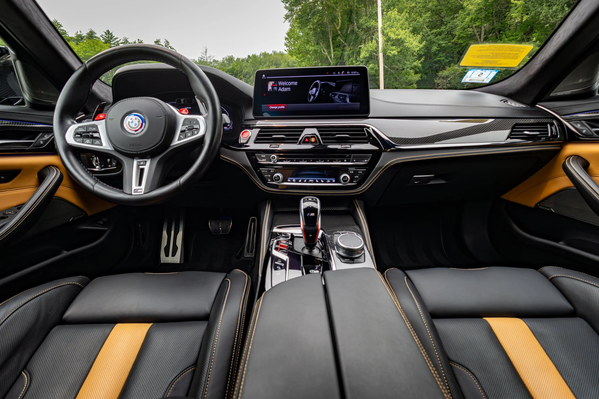 Interior view of a 2024 BMW M5 Competition car dashboard and steering wheel, with a digital display screen showing the name Adam and a welcome message, surrounded by black and tan leather seats with yellow stitching.