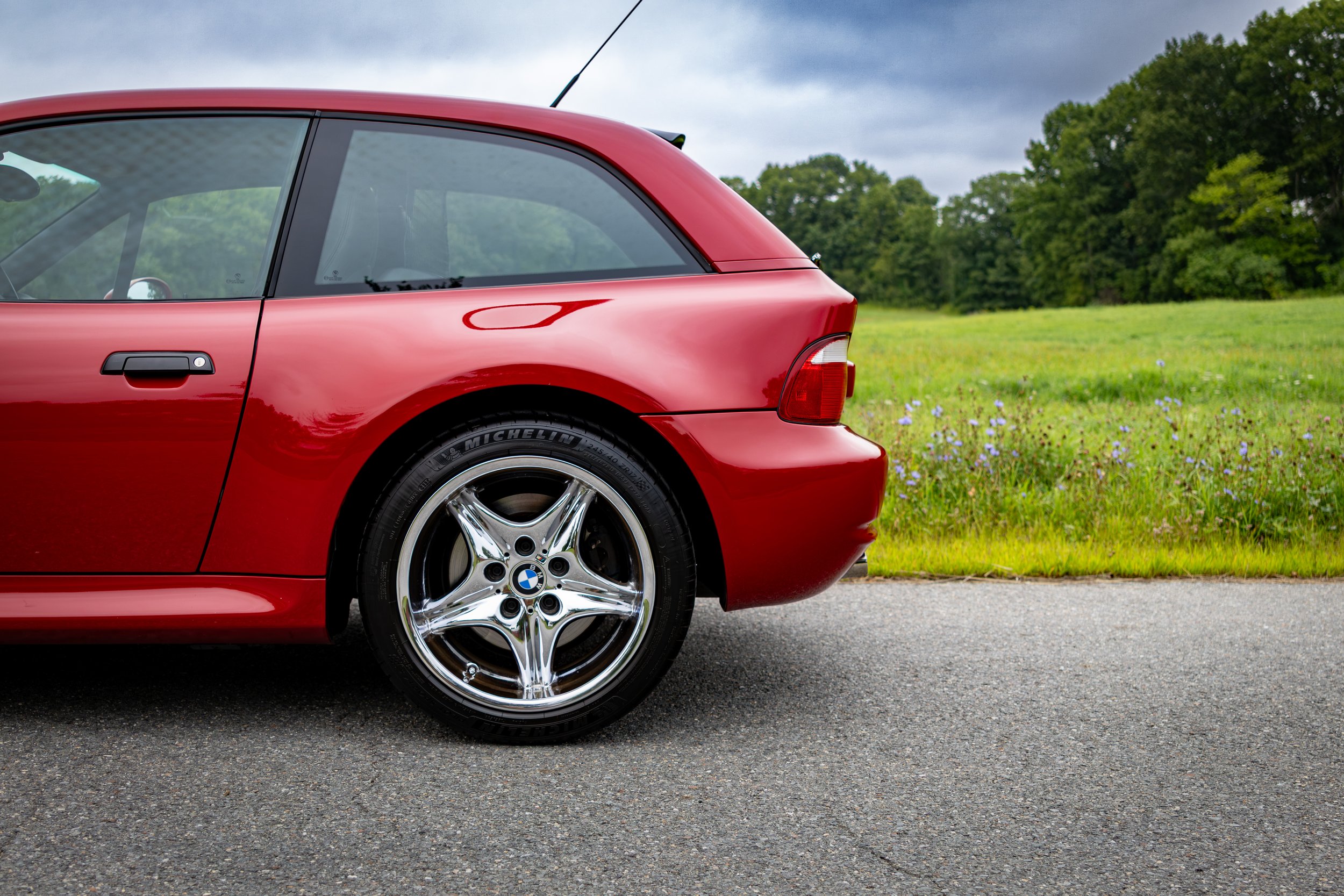 2002 Red BMW M Coupe parked on a road with a grassy field in the background. The rear half of the car is all that is showing from the side.