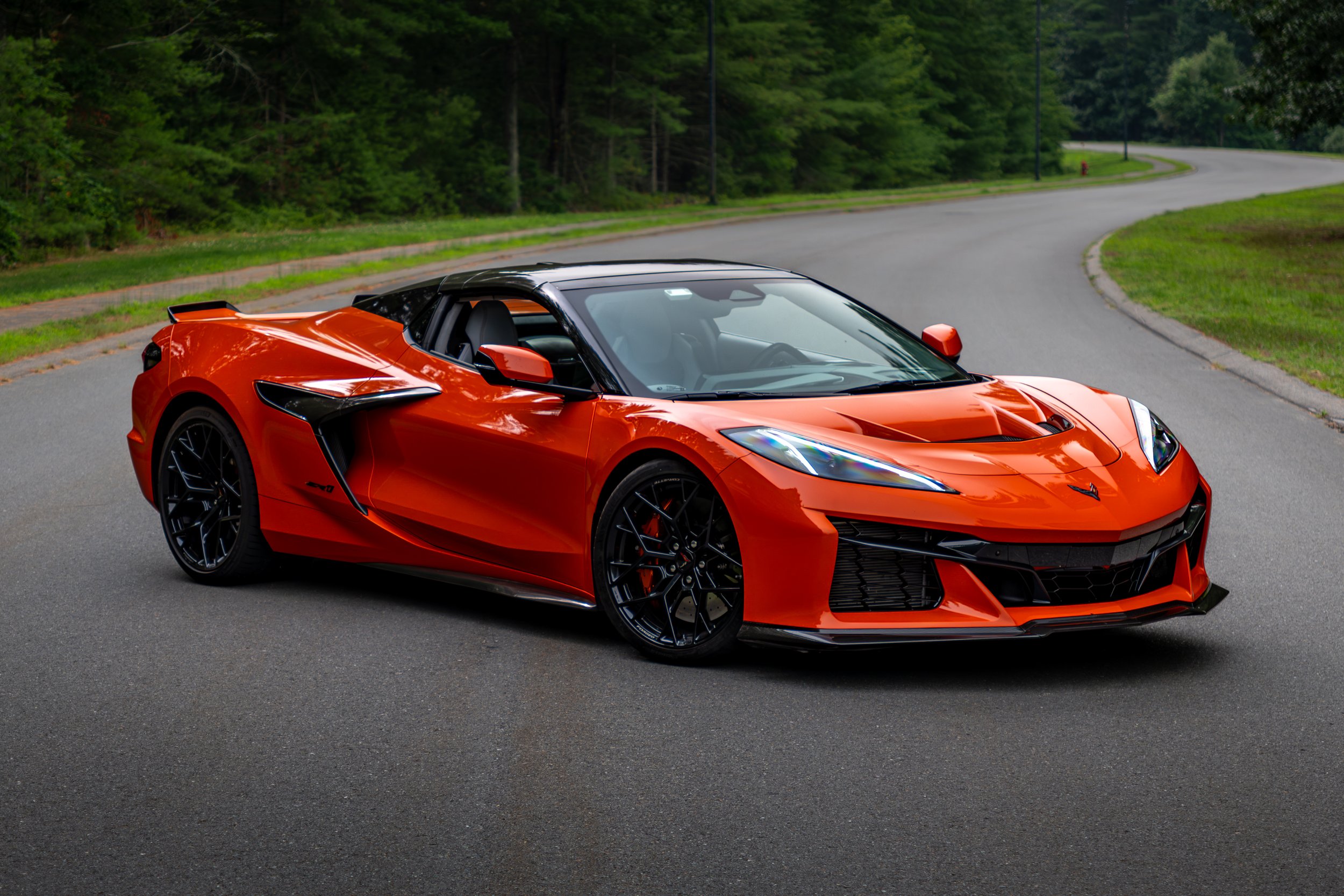 An orange 2026 Chevrolet Corvette ZR1 with 1064 horsepower, parked on a paved road with greenery and trees in the background.