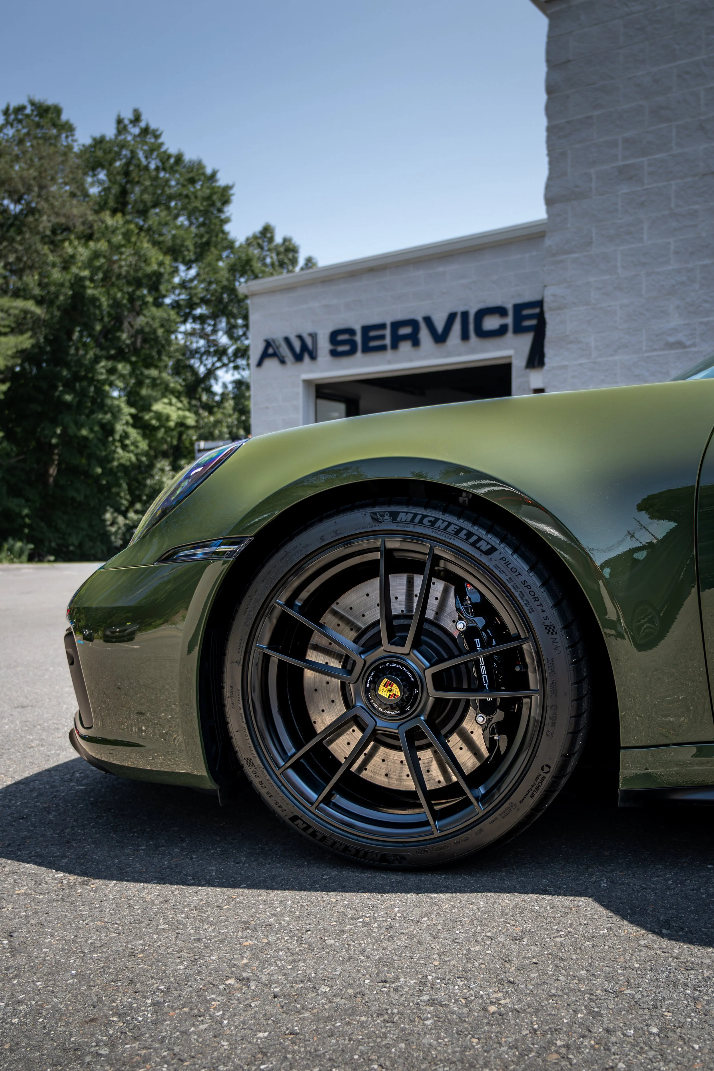Close-up of a nato olive green 2024 Porsche 911 GTS at a Acton Autowerks with a sign that reads "AAW SERVICE" on the building in the background, trees, and a clear blue sky.
