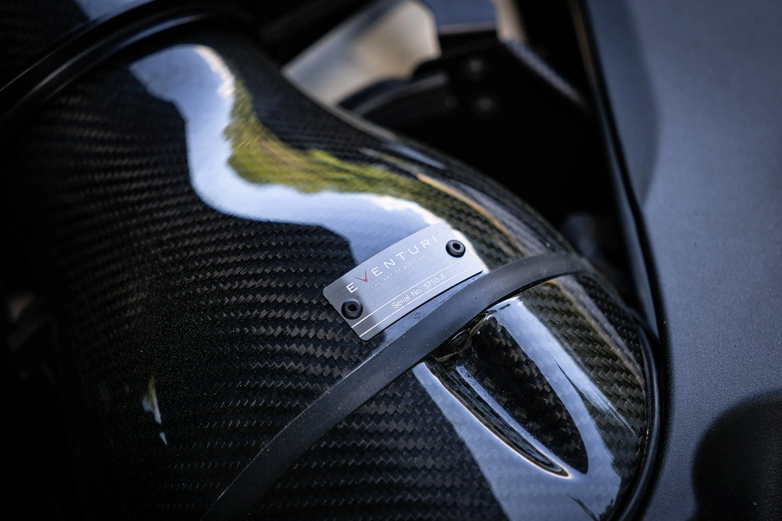 Close-up of an Eventuri carbon fiber intake on a flat black BMW M5 Competition with a metal brand emblem that reads 'EVENTURI The Art of Airflow.'