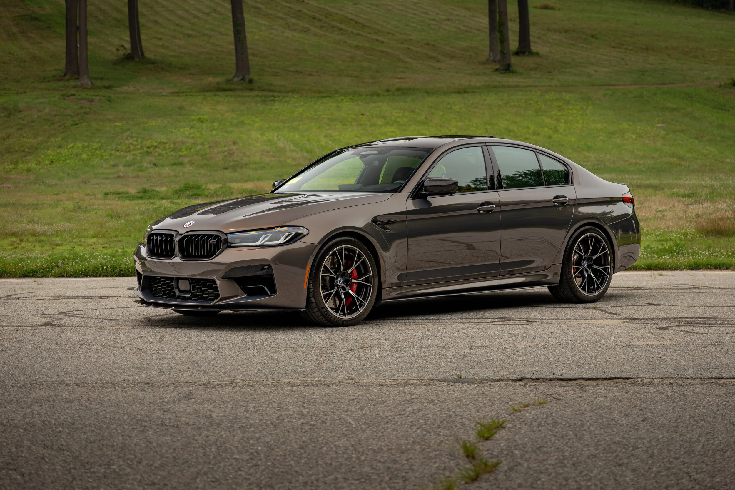 A sleek brown BMW M5 Competitions parked on a paved surface with a grassy field and trees in the background.