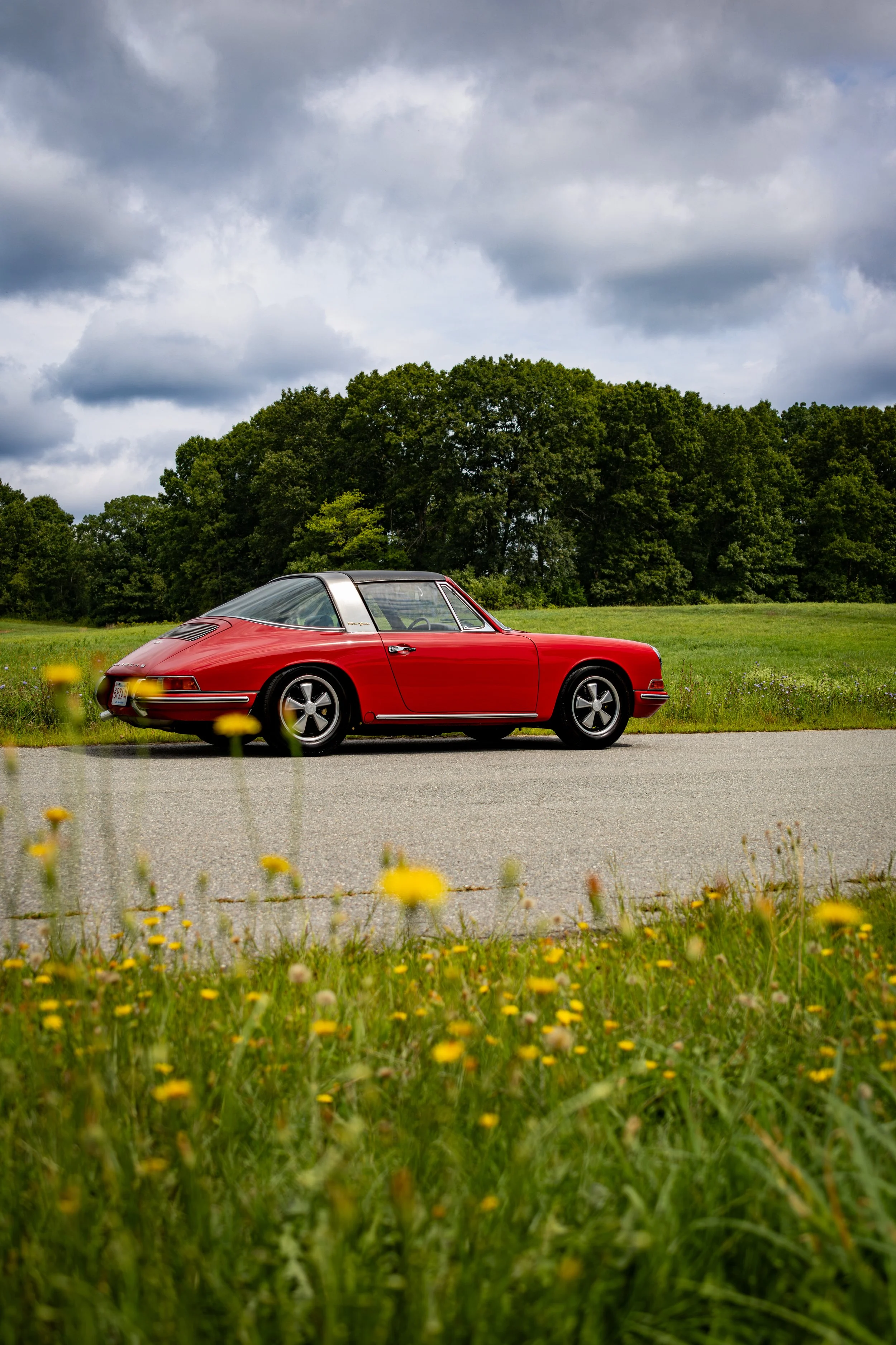 1968 Guards Red Porsche 911 Targa parked on a road in a grassy field. Yellow flowers are sitting in the foreground of the photo but out of focus. 