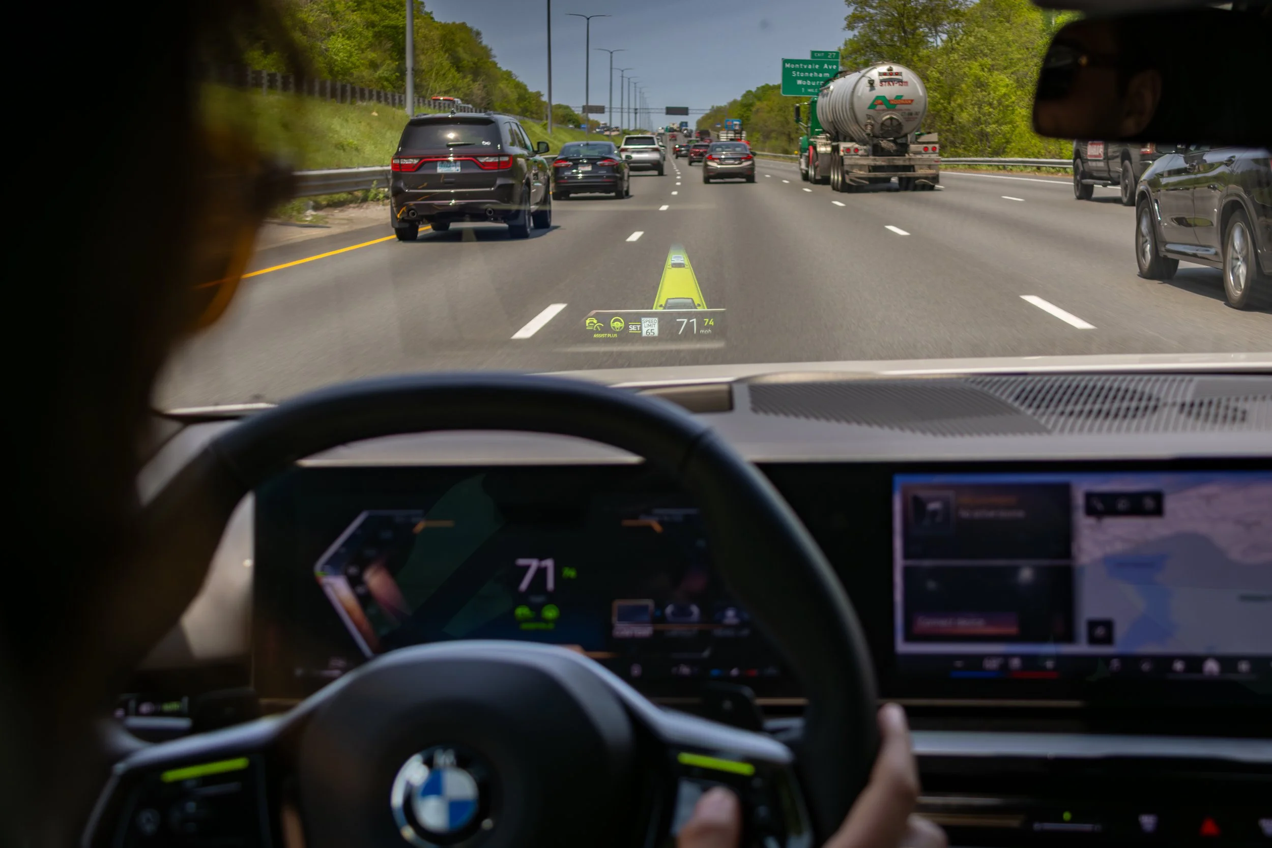 A view from inside a MIT Age Lab, BMW 5-series test vehicle on a busy multi-lane highway with cars and trucks, some trees on the roadside, and highway signs in the background. Heads-up display is visible