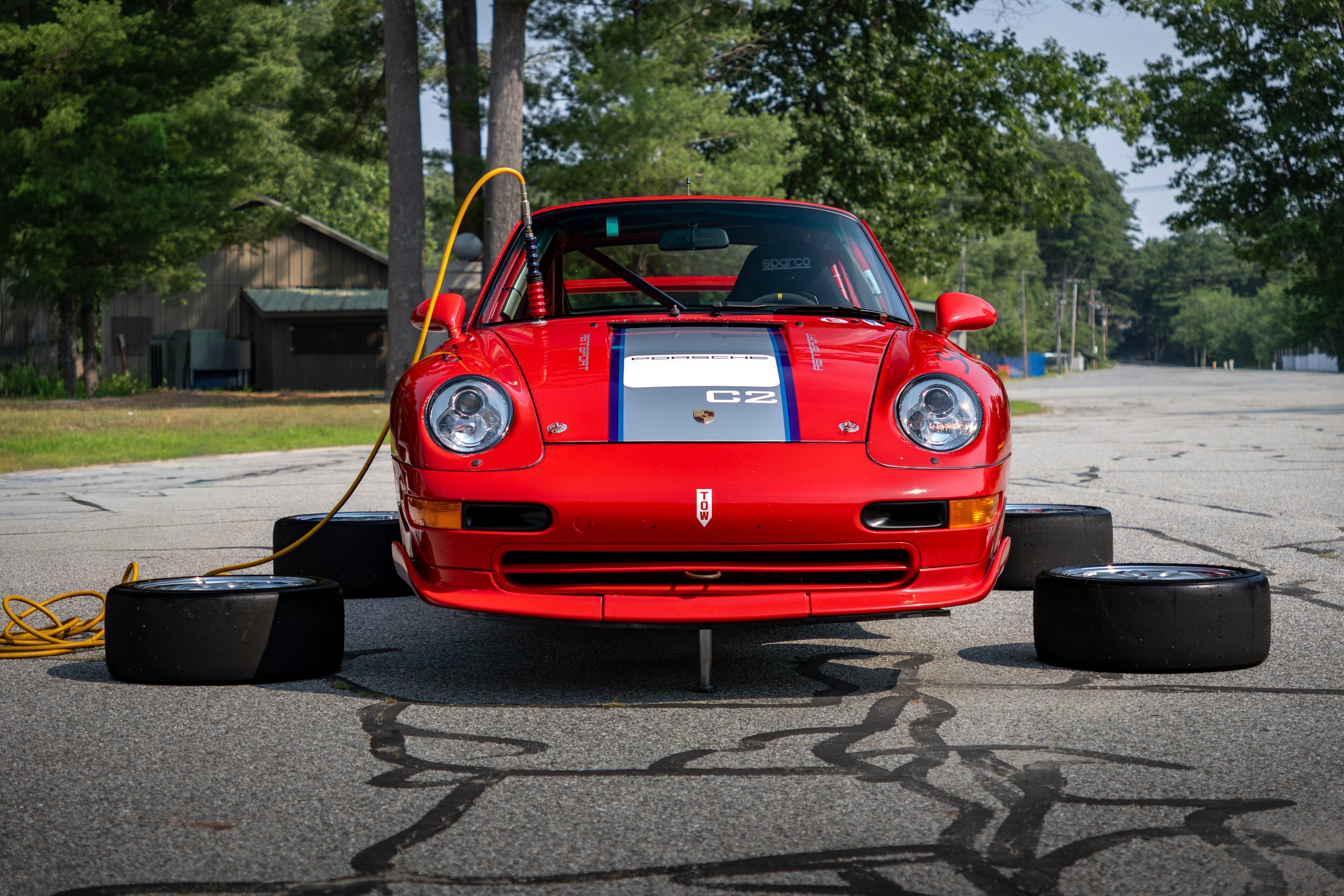 A red Porsche GT2 Supercup race car with a silver and blue racing stripe on the hood, parked on a cracked asphalt surface with tires and a yellow power cable nearby, surrounded by trees.