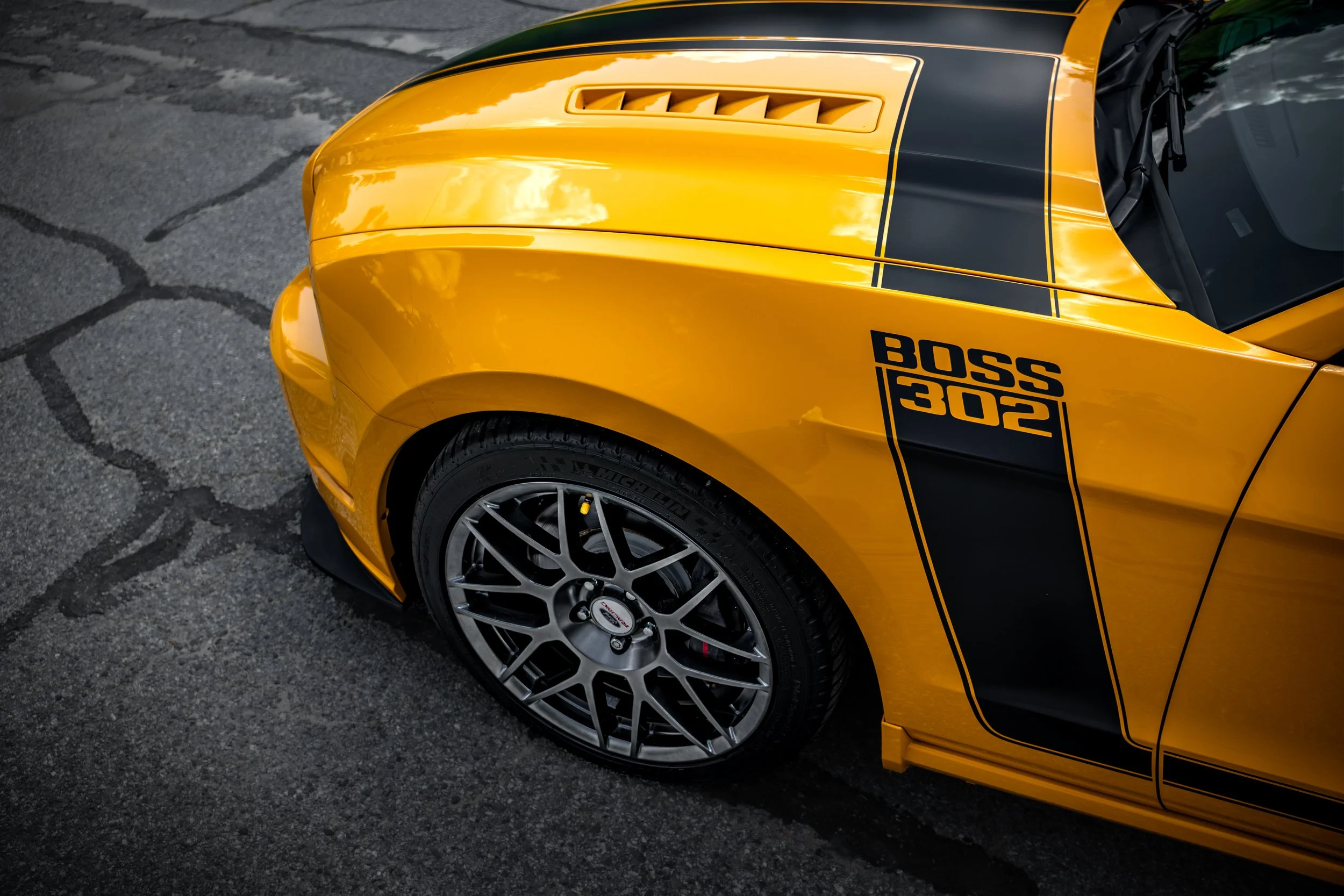 Close-up of a yellow sports car with black racing stripes and a vented hood, parked on asphalt.
