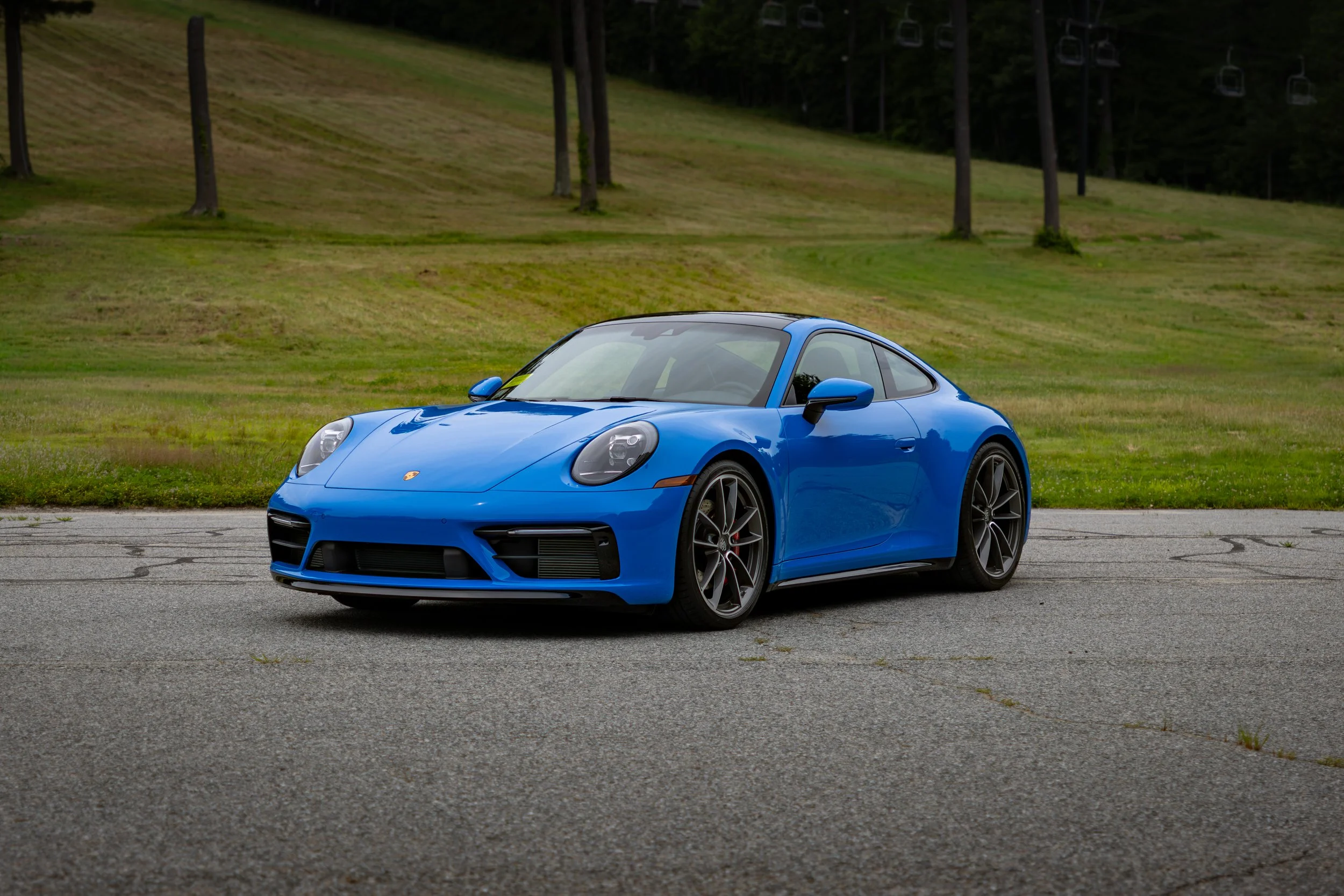 A shark blue 2024 Porsche 911 Carrera S parked on a paved surface with a grassy hill and trees in the background.