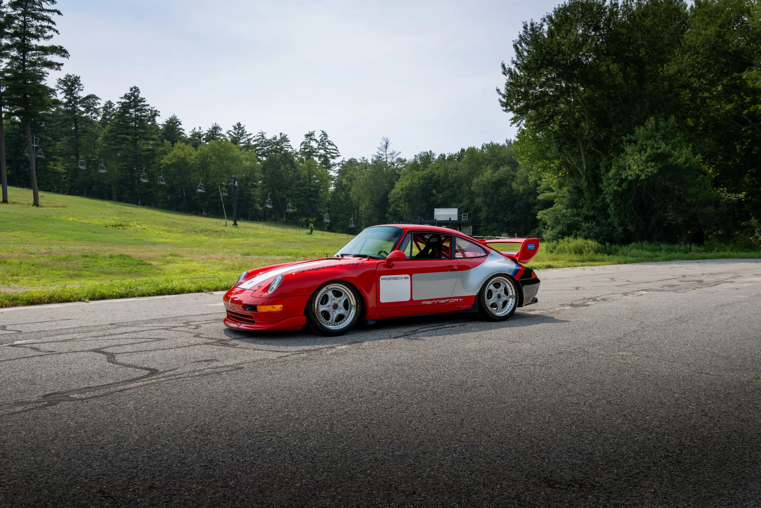 A red and white Porsche race car parked on a cracked asphalt road with green trees, a grassy area, and ski lift chairs in the background.