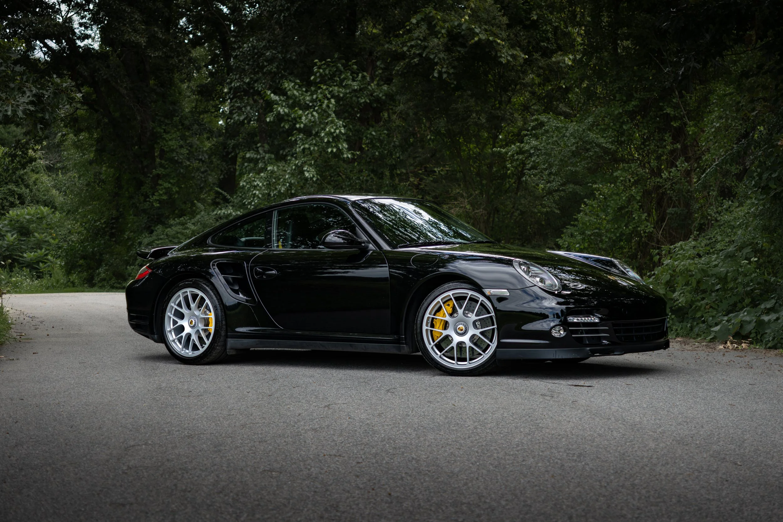 2019 Porsche 911 Turbo S (997) in Black with Carbon Ceramic brakes (PCCBs) parked on a road with nothing but greenery and trees in the background.