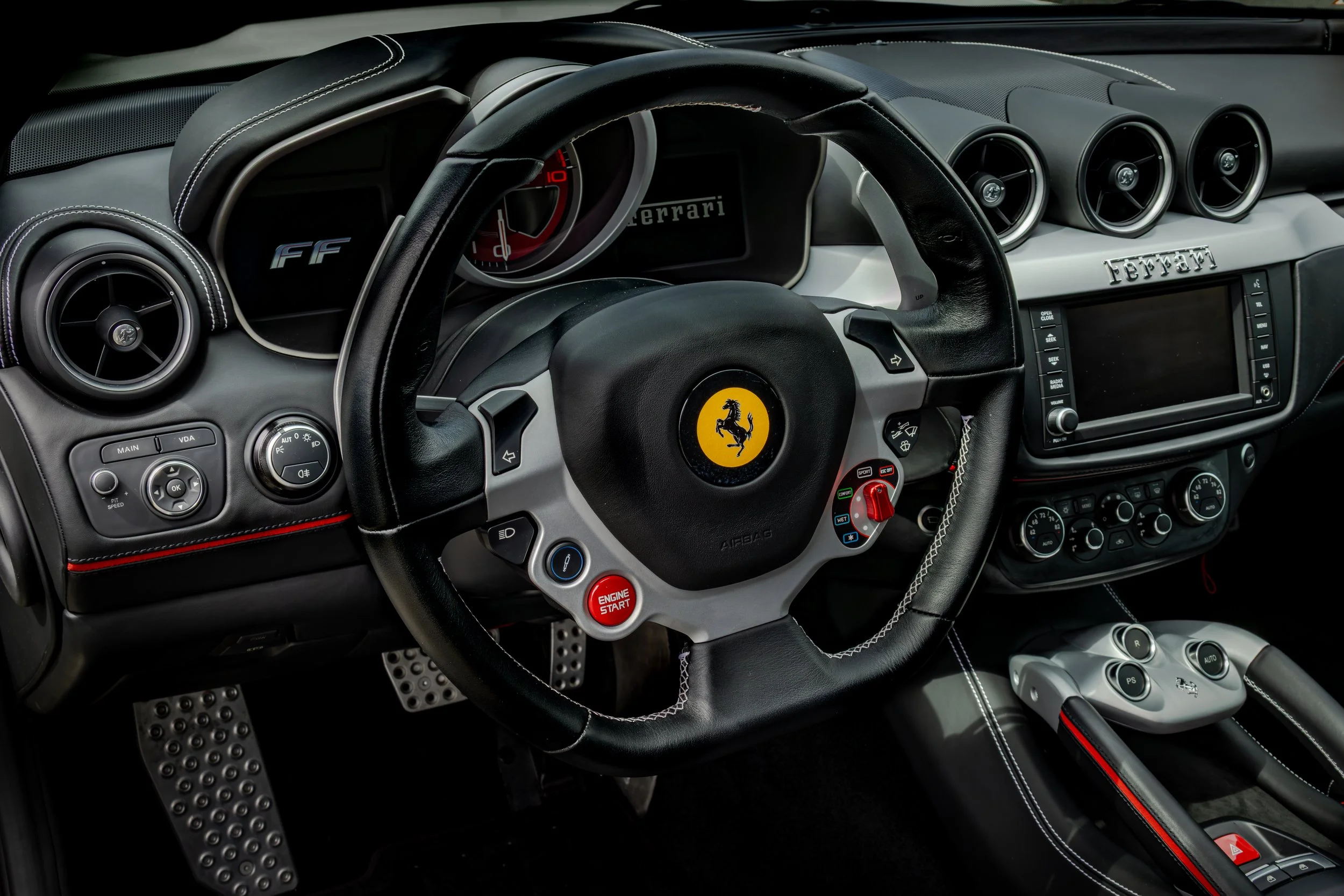 The interior of a Ferrari FF, featuring a black leather steering wheel with the yellow Ferrari logo, a digital dashboard with red accents, three circular air vents, and various control buttons and dials on the dashboard and center console.