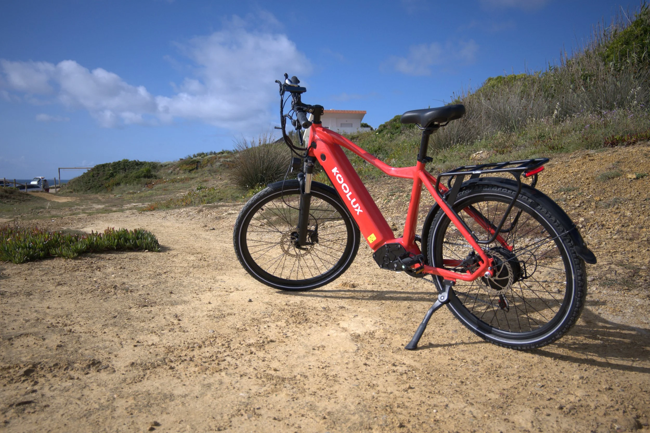 Red electric bicycle parked on a dirt path near shrubs with a small white house and blue sky with clouds in the background.