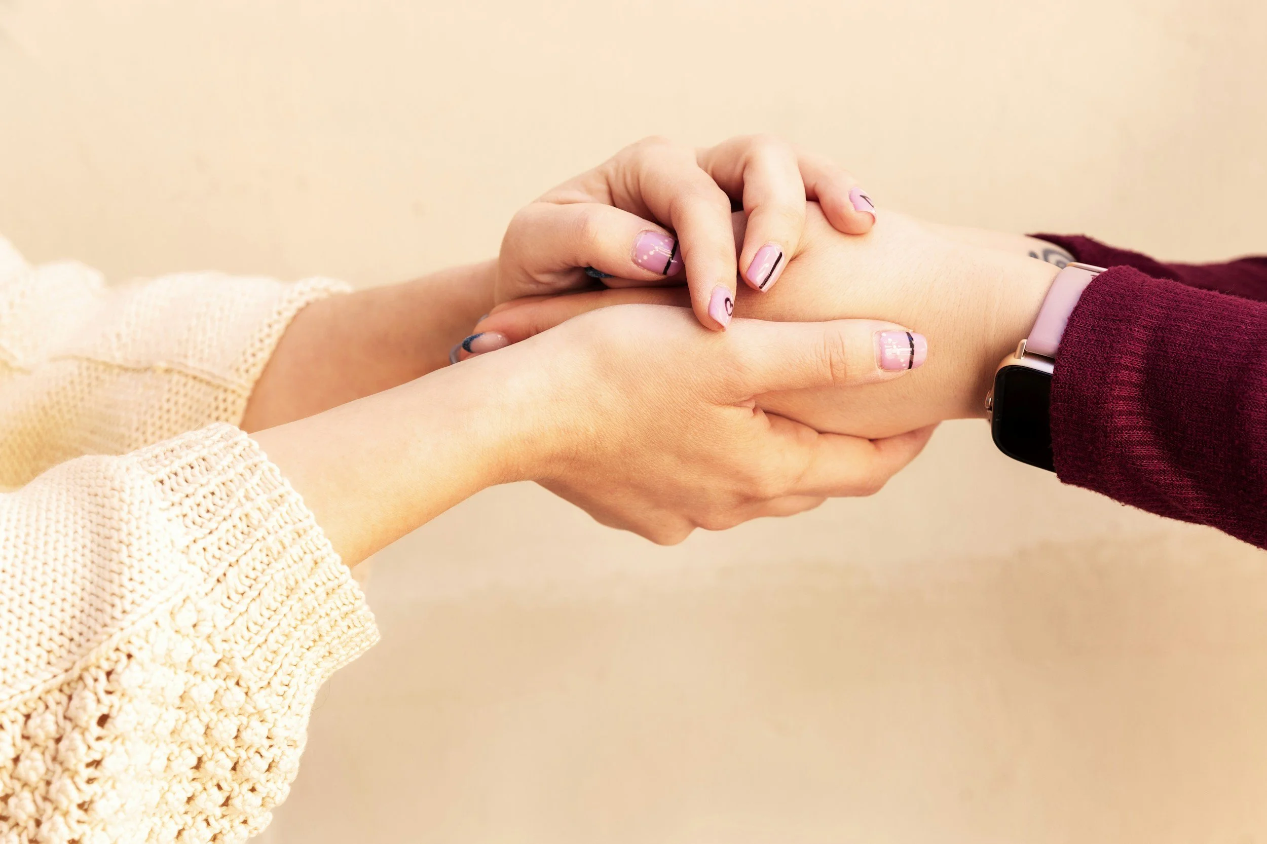 Two people holding hands, one with a pink smartwatch and pink and black nail polish, and the other with a yellow knit sweater. The background is plain off-white.
