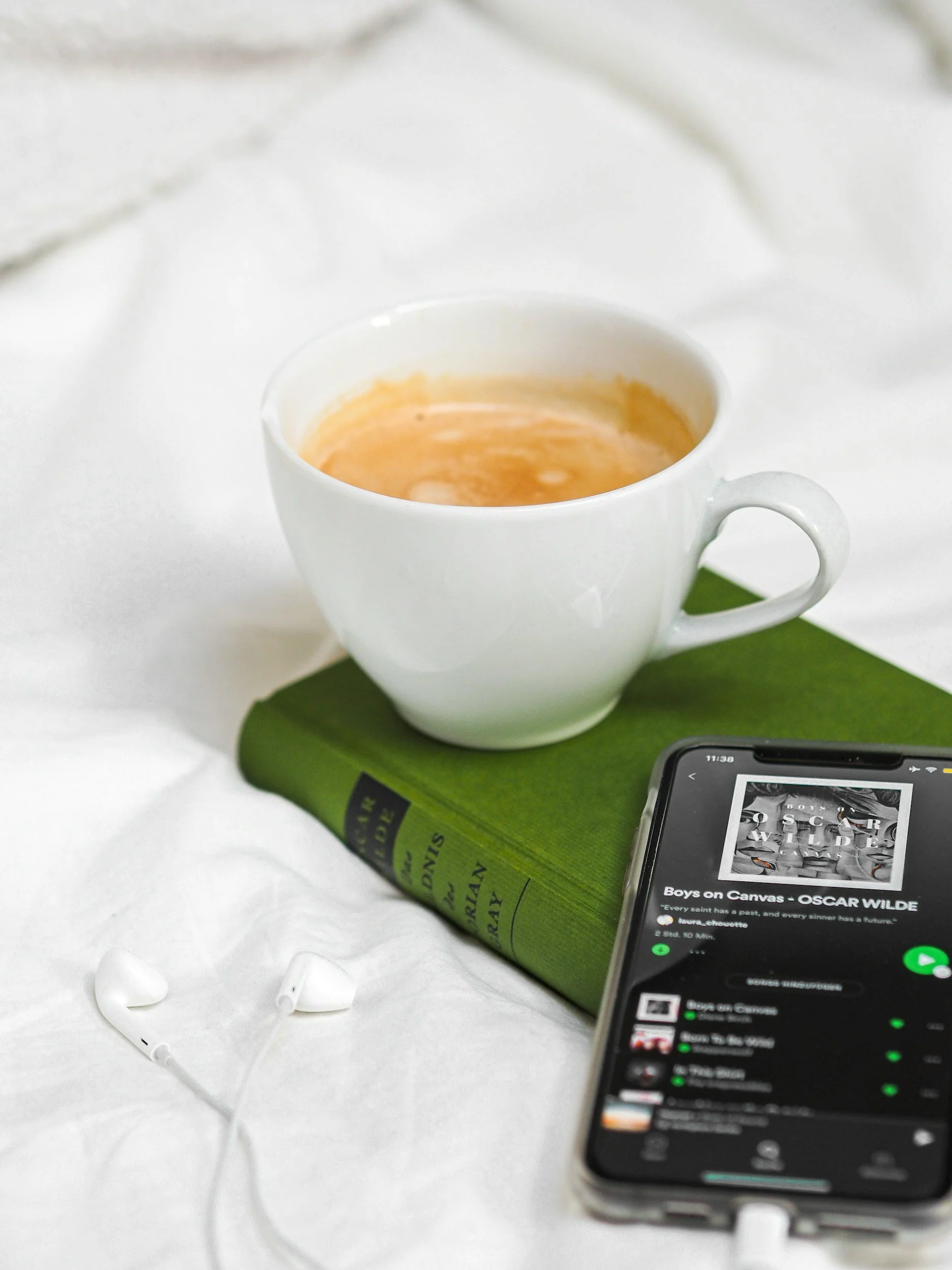 A white ceramic mug filled with coffee on a green book, with white earphones and a smartphone displaying a music playlist on a white surface.