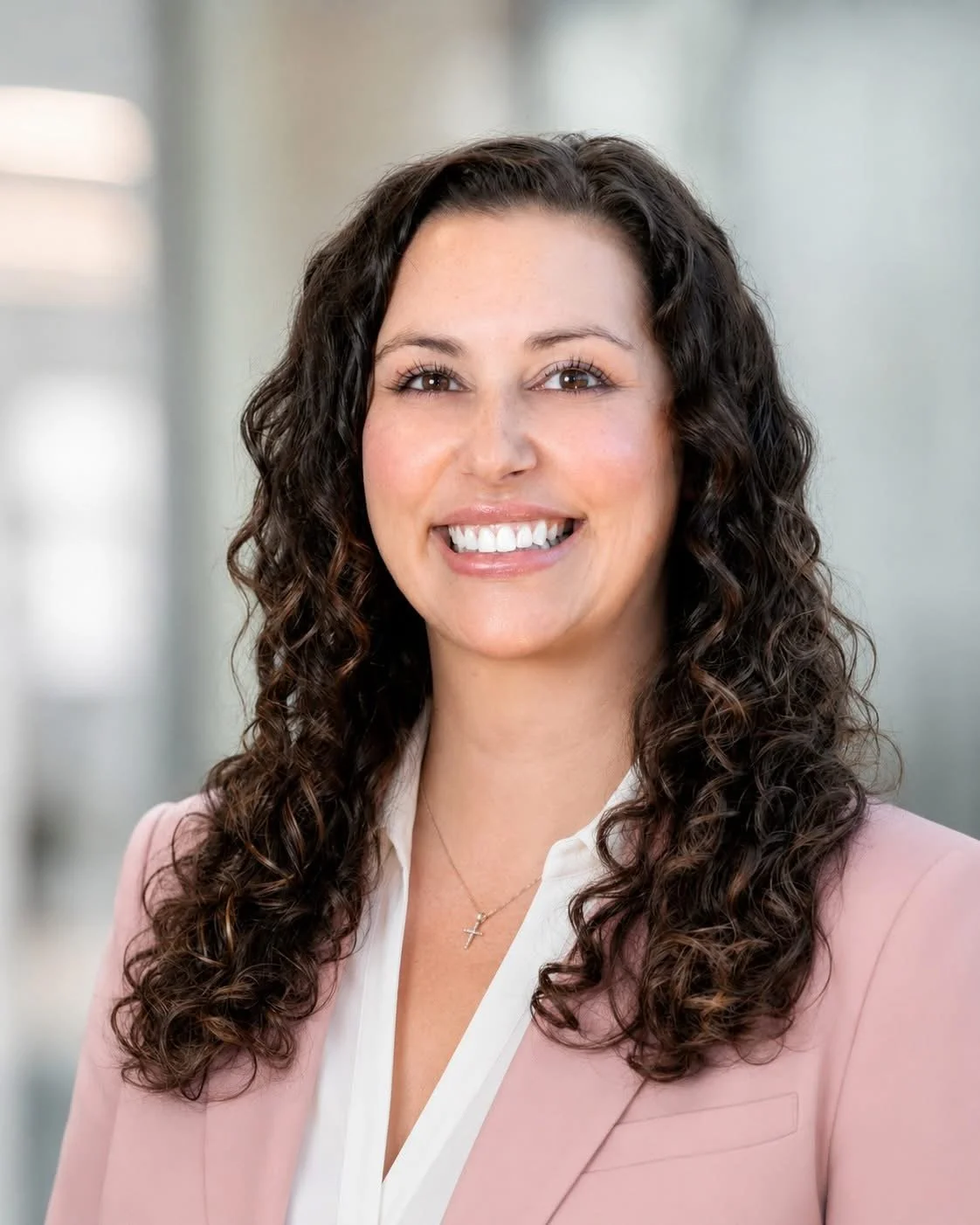 Professional woman with long, curly dark hair, smiling, wearing a pink blazer and a white blouse, in a bright office setting.