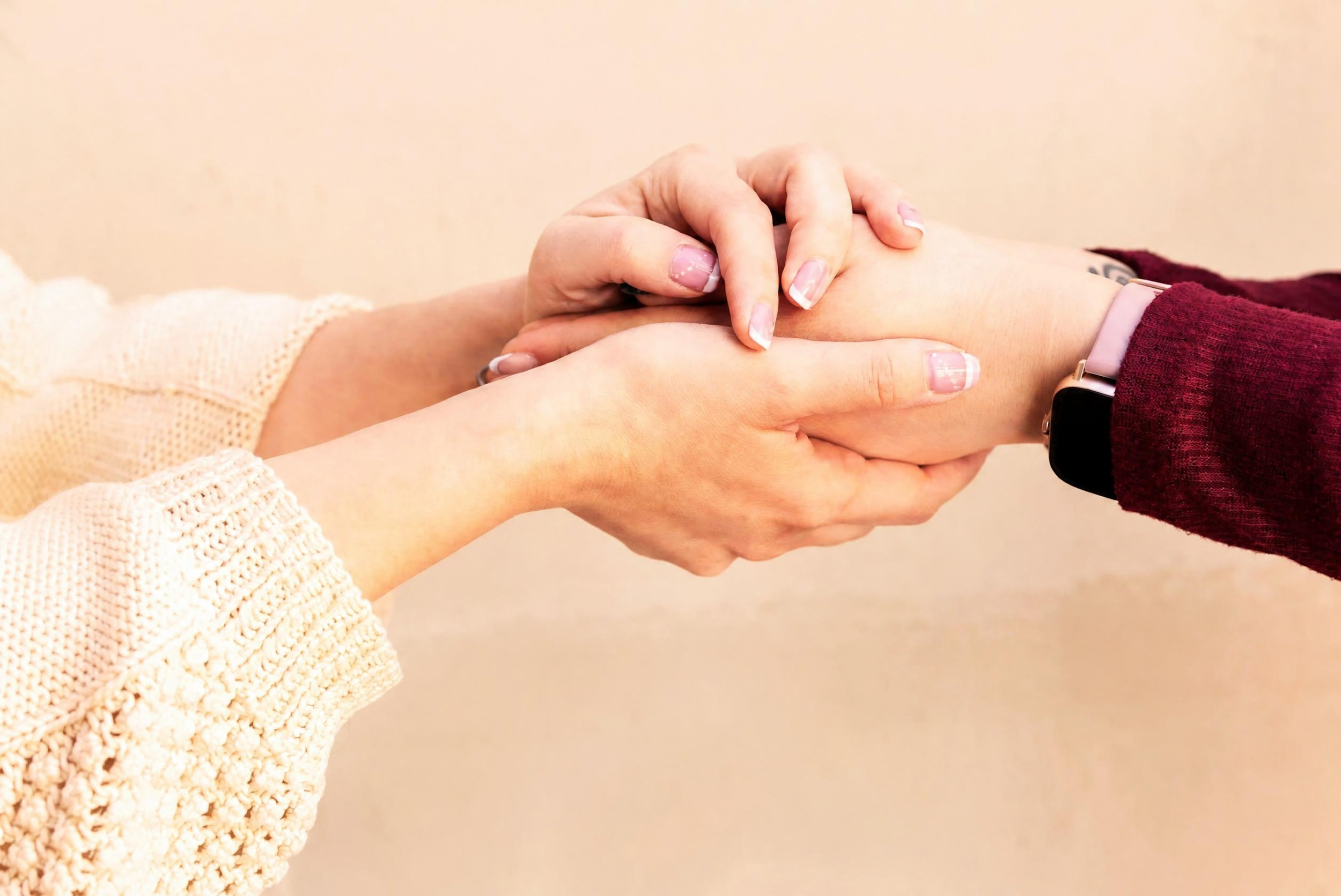 Close-up of two people holding hands, one with a smartwatch, wearing a maroon sweater, and the other with manicured nails, wearing a cream sweater.
