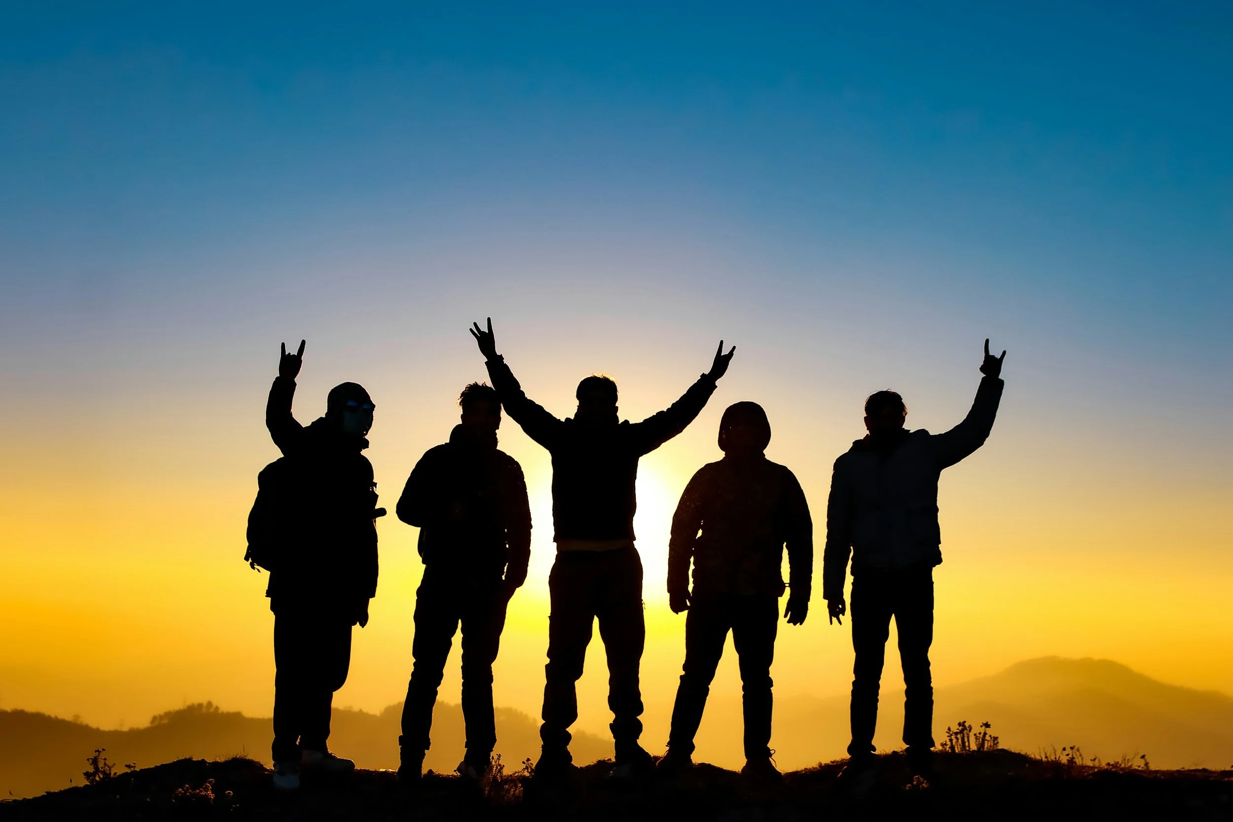 Silhouettes of five people standing together outdoors at sunset or sunrise, making peace signs and raising their hands, with a clear sky and distant hills in the background.