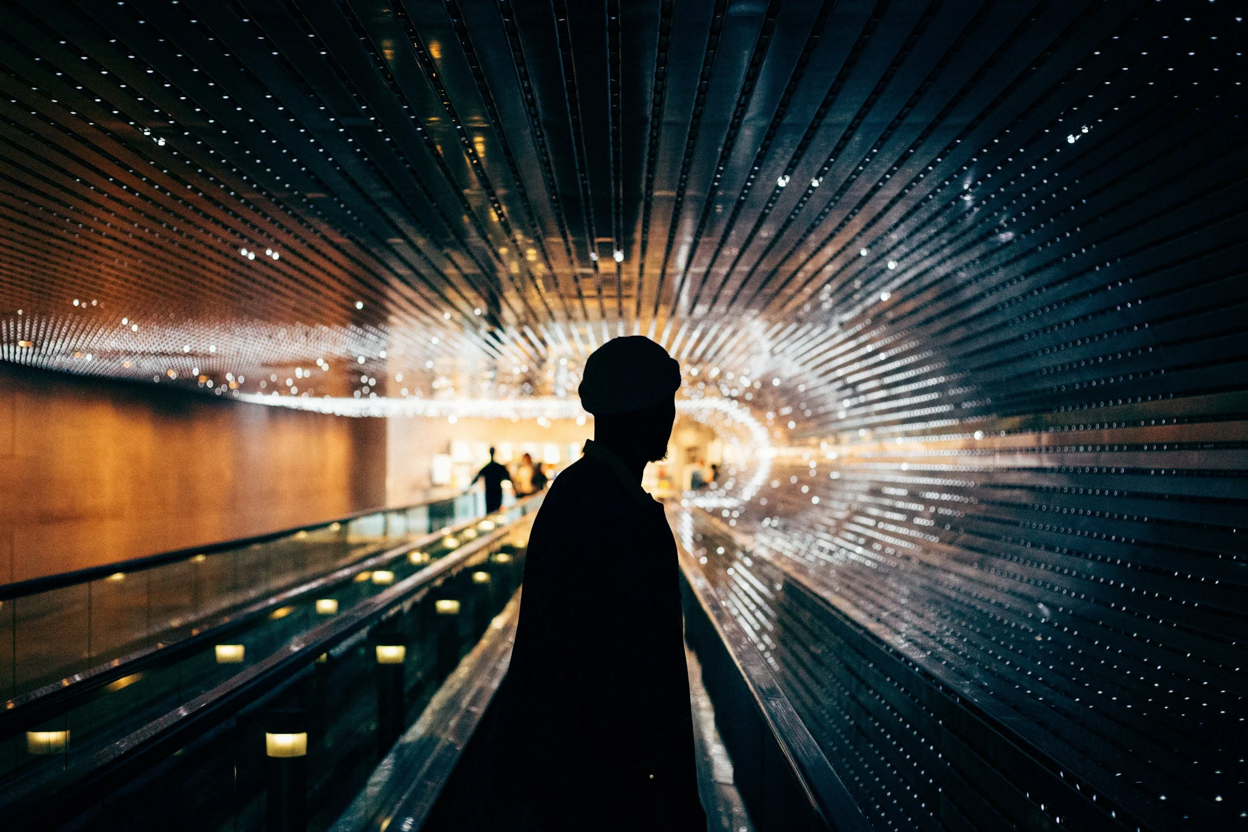 Silhouette of a person standing in an illuminated tunnel-like structure with circular light patterns on the ceiling.