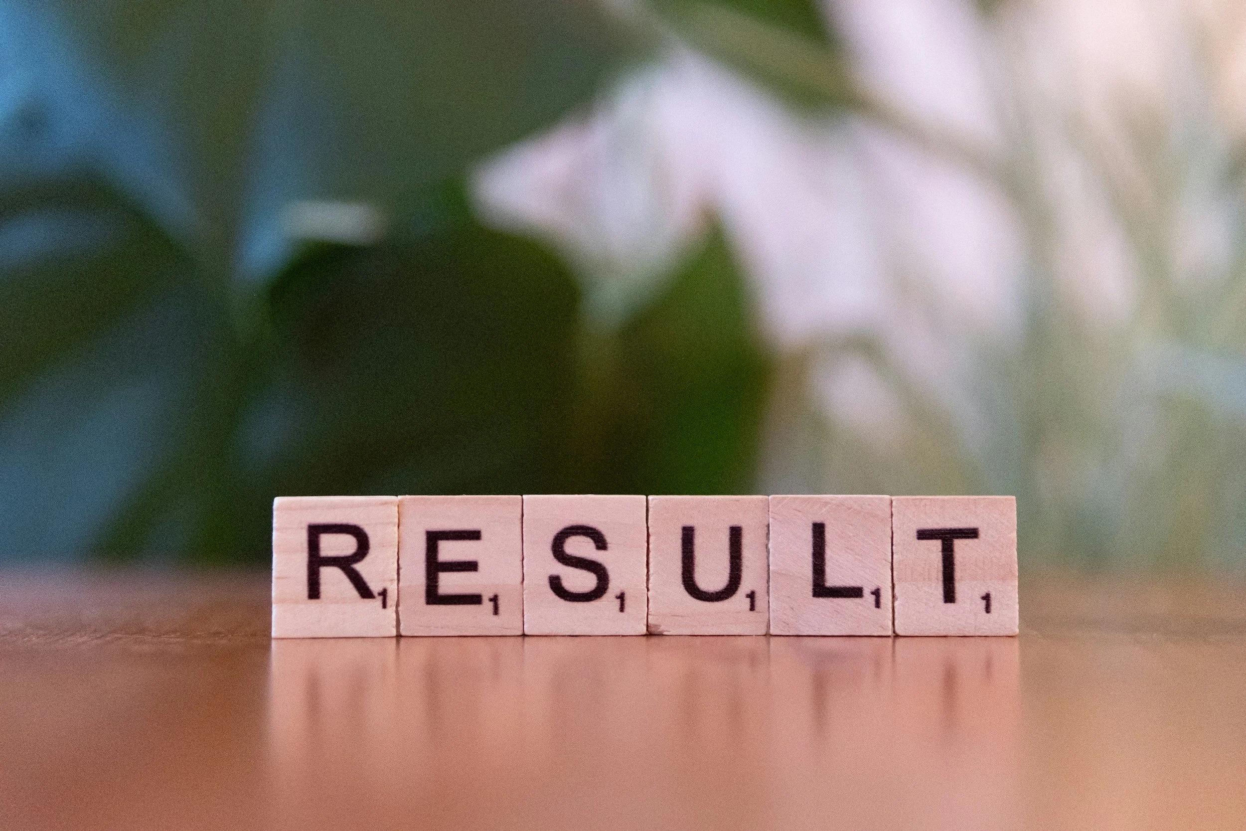 Bloques de letras que forman la palabra "RESULT" en una superficie de madera, con fondo borroso de tonos verdes y blancos.