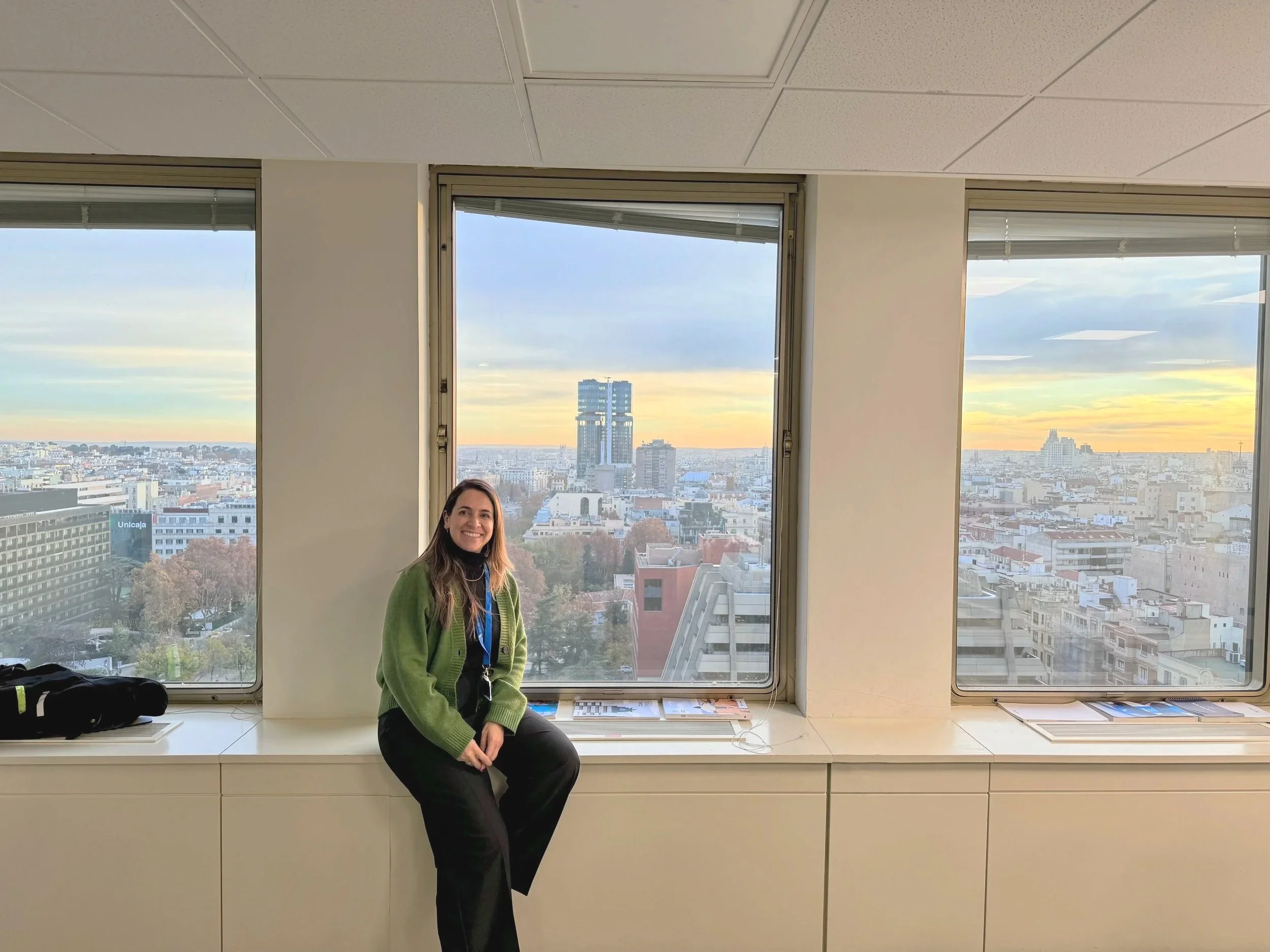 Una mujer sonriente de cabello largo y oscuro, usando un suéter verde y pantalones negros, sentada en un escritorio frente a una ventana grande con vista a la ciudad al atardecer o amanecer.