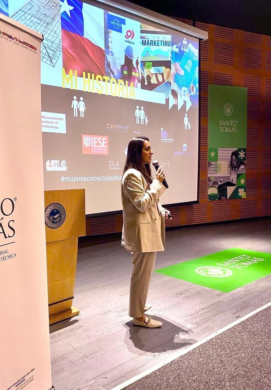 Mujer joven de espaldas, con traje beige y sandalias, hablando con micrófono en una sala de conferencias, con pantalla grande detrás que muestra una presentación titulada 'Mi Historia' y logotipos de varias instituciones educativas, en un entorno interior con paredes de madera y señalización verde de Santo Tomás.