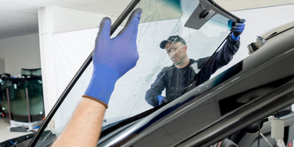 Two people wearing gloves, one is removing or inspecting a cracked windshield of a vehicle in a garage or repair shop.