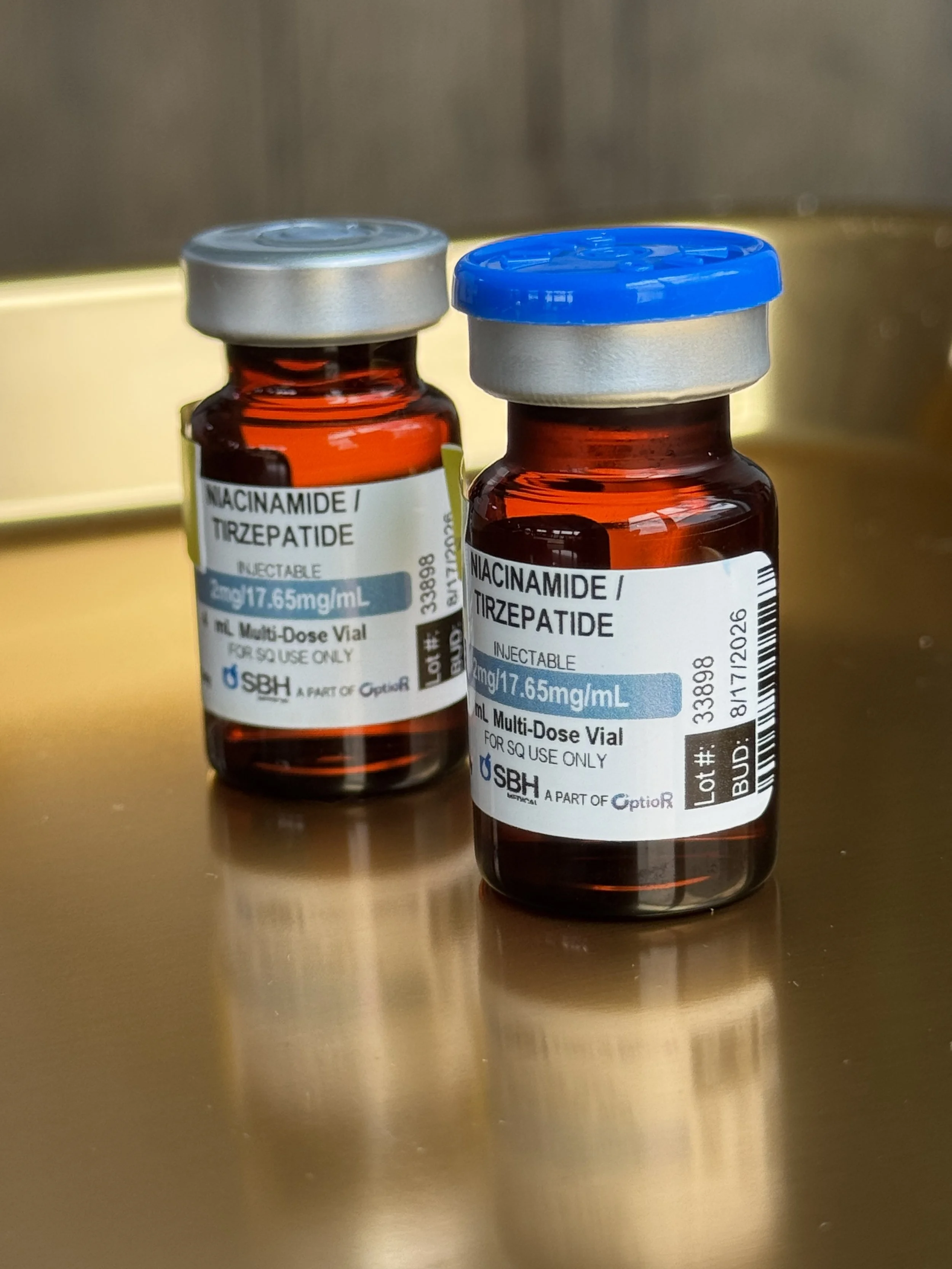Two small pharmacy vials labeled with medication information, one with a silver cap and the other with a blue cap, placed on a shiny gold-colored surface.