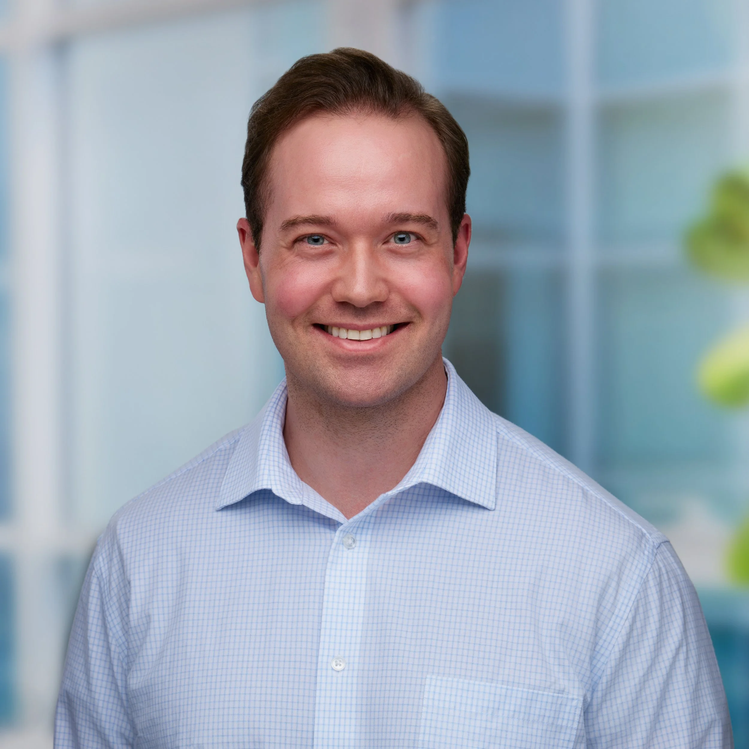 Close-up of a smiling man wearing a light blue checkered shirt, with a blurred office background.