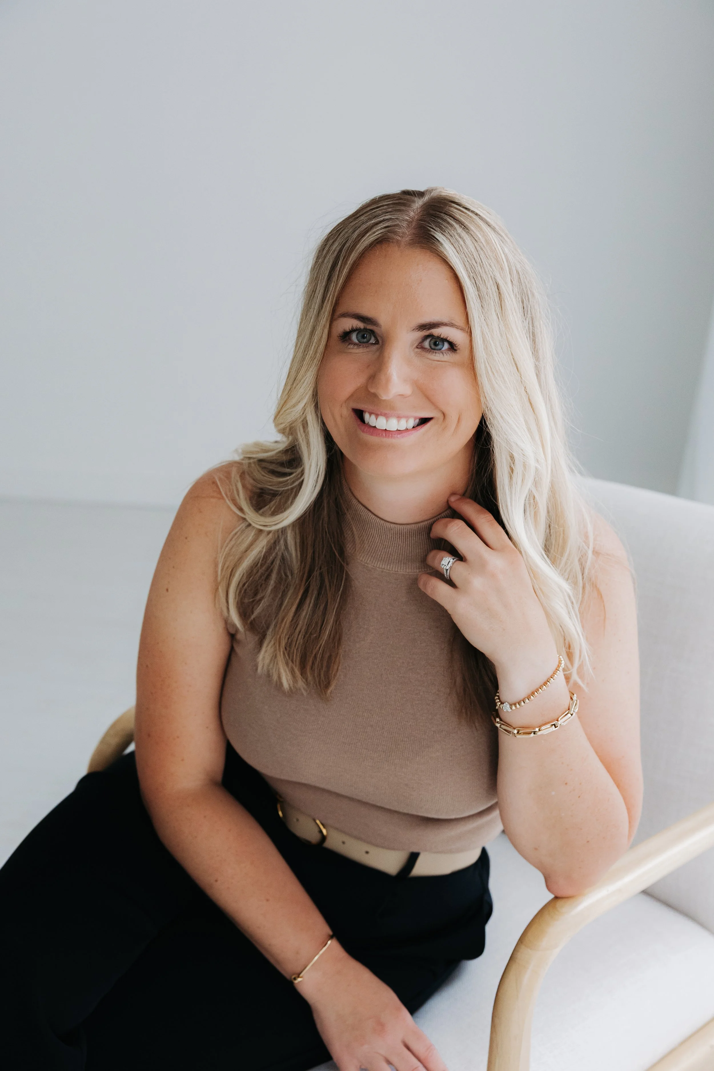 A woman with long blonde hair, smiling, sitting on a white chair with a curved wooden armrest. She is wearing a beige sleeveless top, black pants, and gold jewelry, including a bracelet, rings, and a watch.