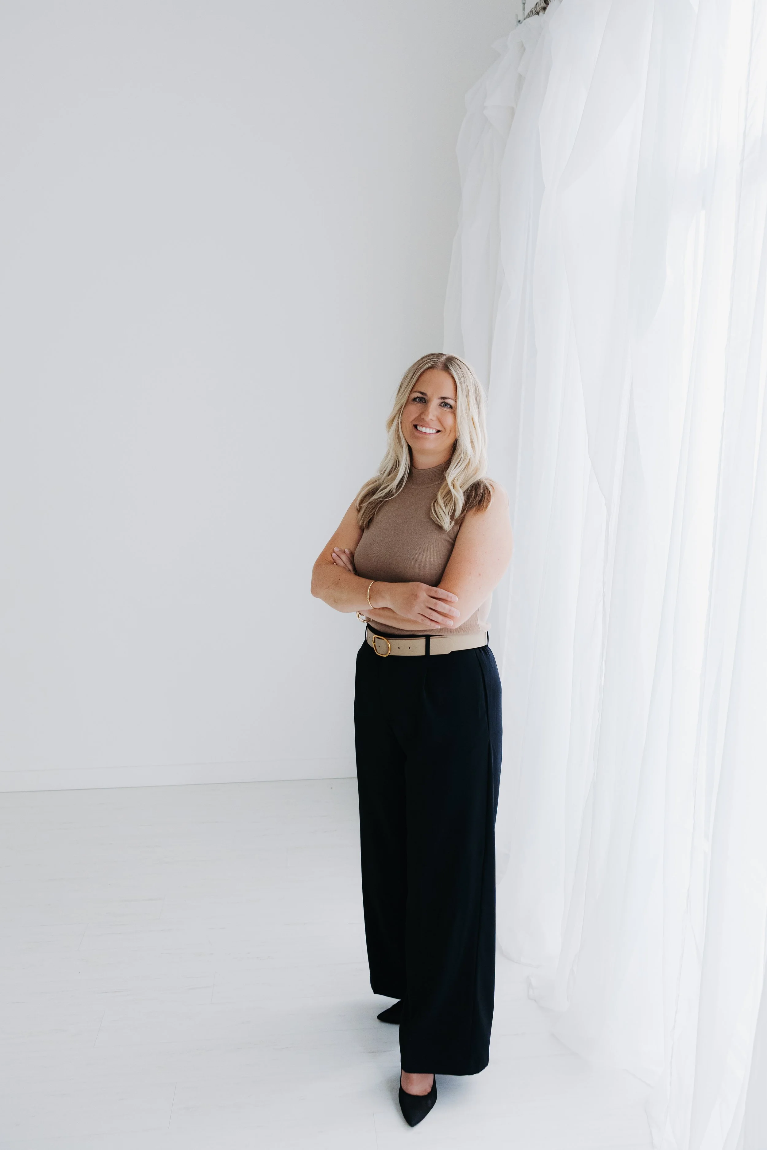 A woman with blonde hair stands with arms crossed, smiling, in front of a white wall and sheer white curtains.