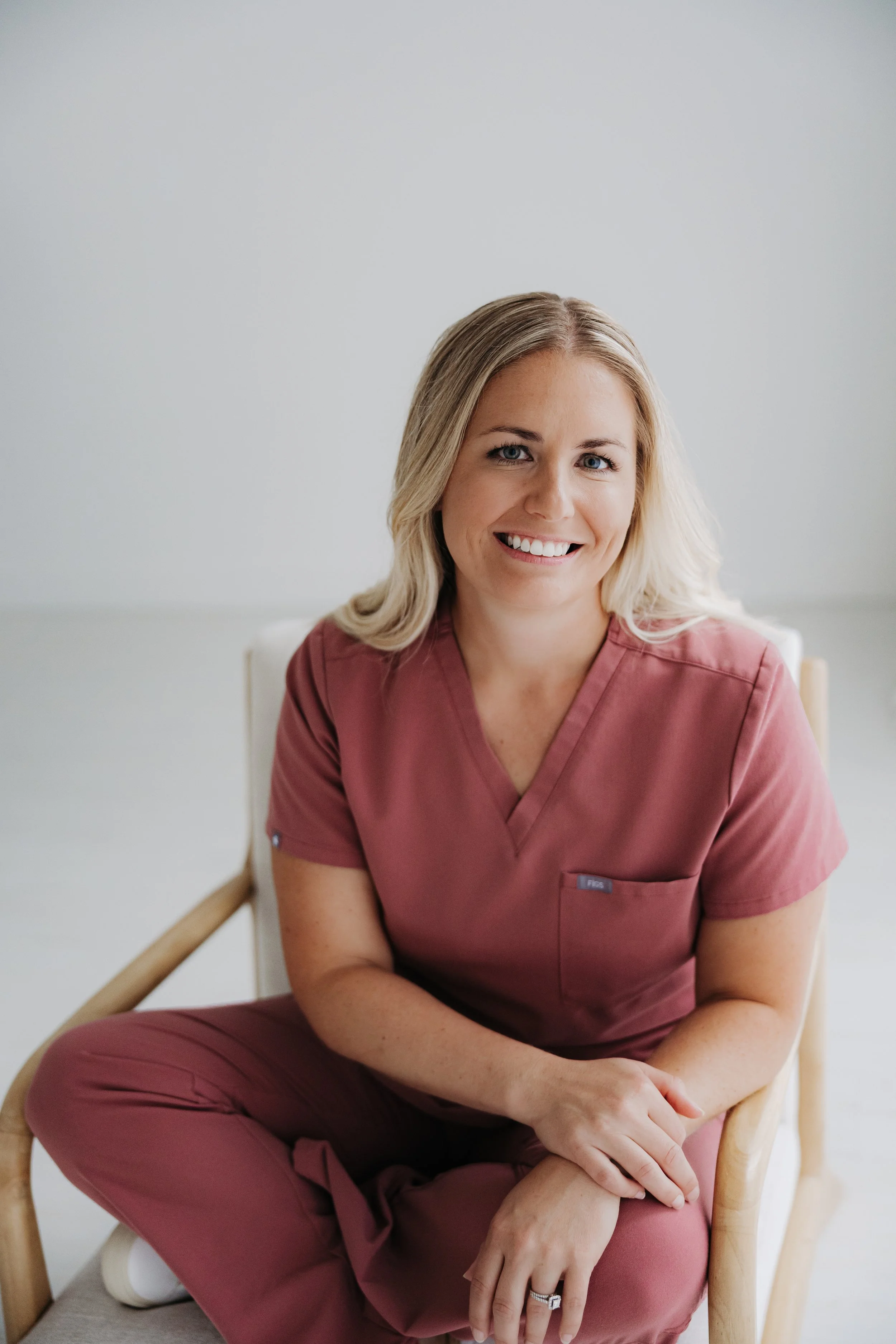 A smiling woman with blonde hair wearing pink medical scrubs, sitting on a wooden chair with armrests in a room with plain light-colored walls.
