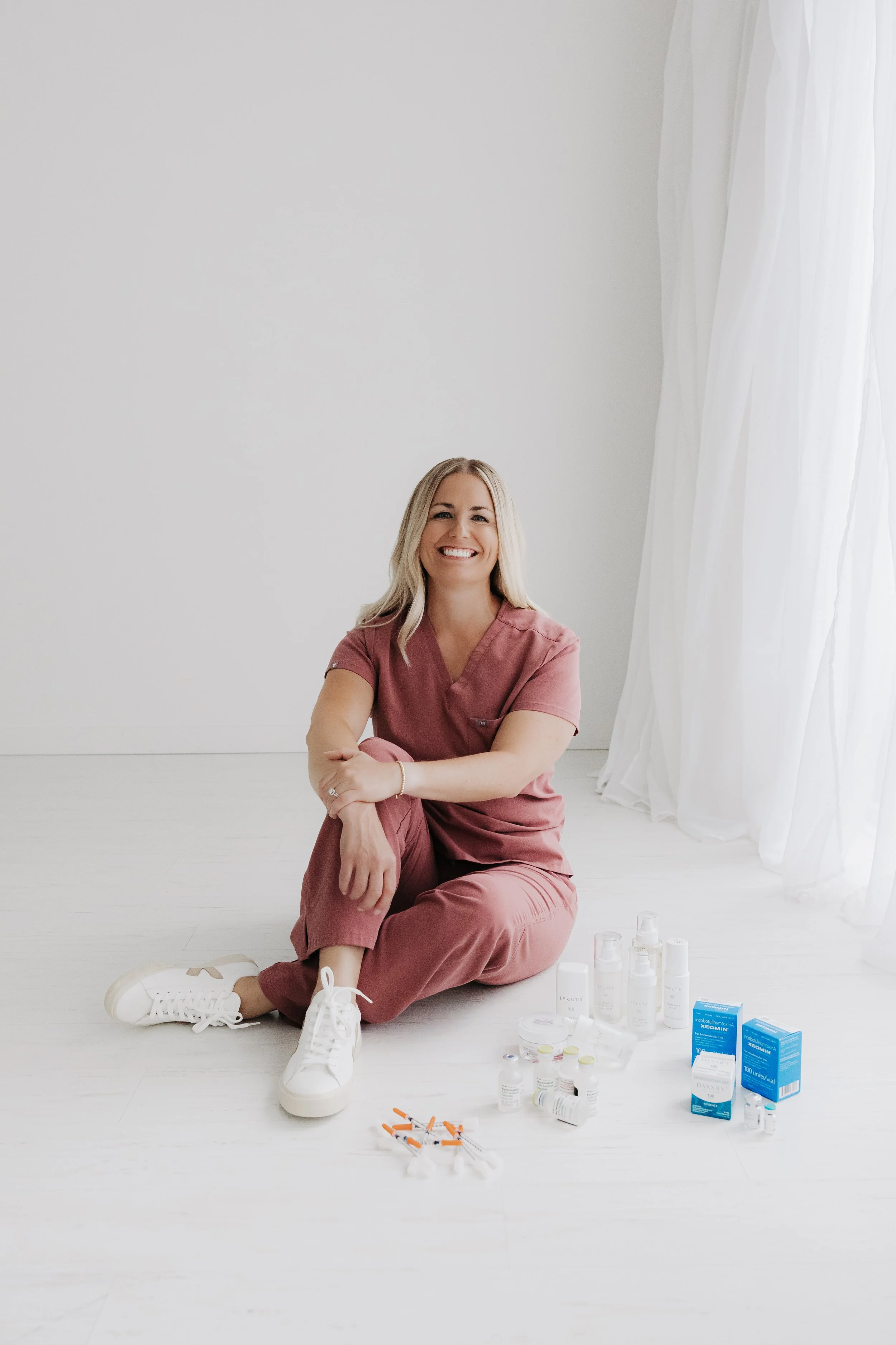 A smiling woman in pink scrubs sitting on the floor with her legs crossed, surrounded by medical supplies and medications, in a bright room with white walls and curtains.