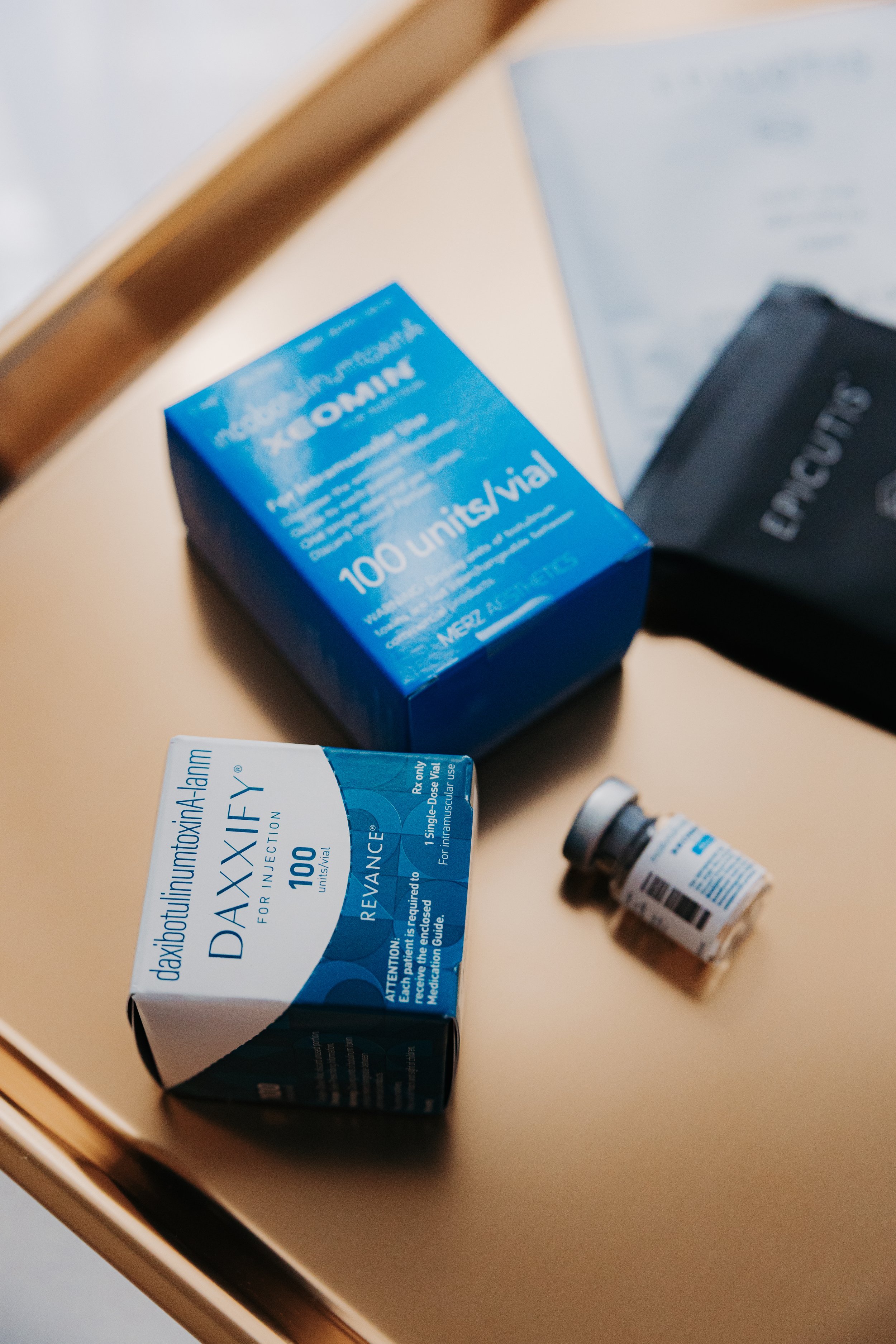 Boxes of Daxify and Xeomin neuromodulators and a small vial on a gold surface.