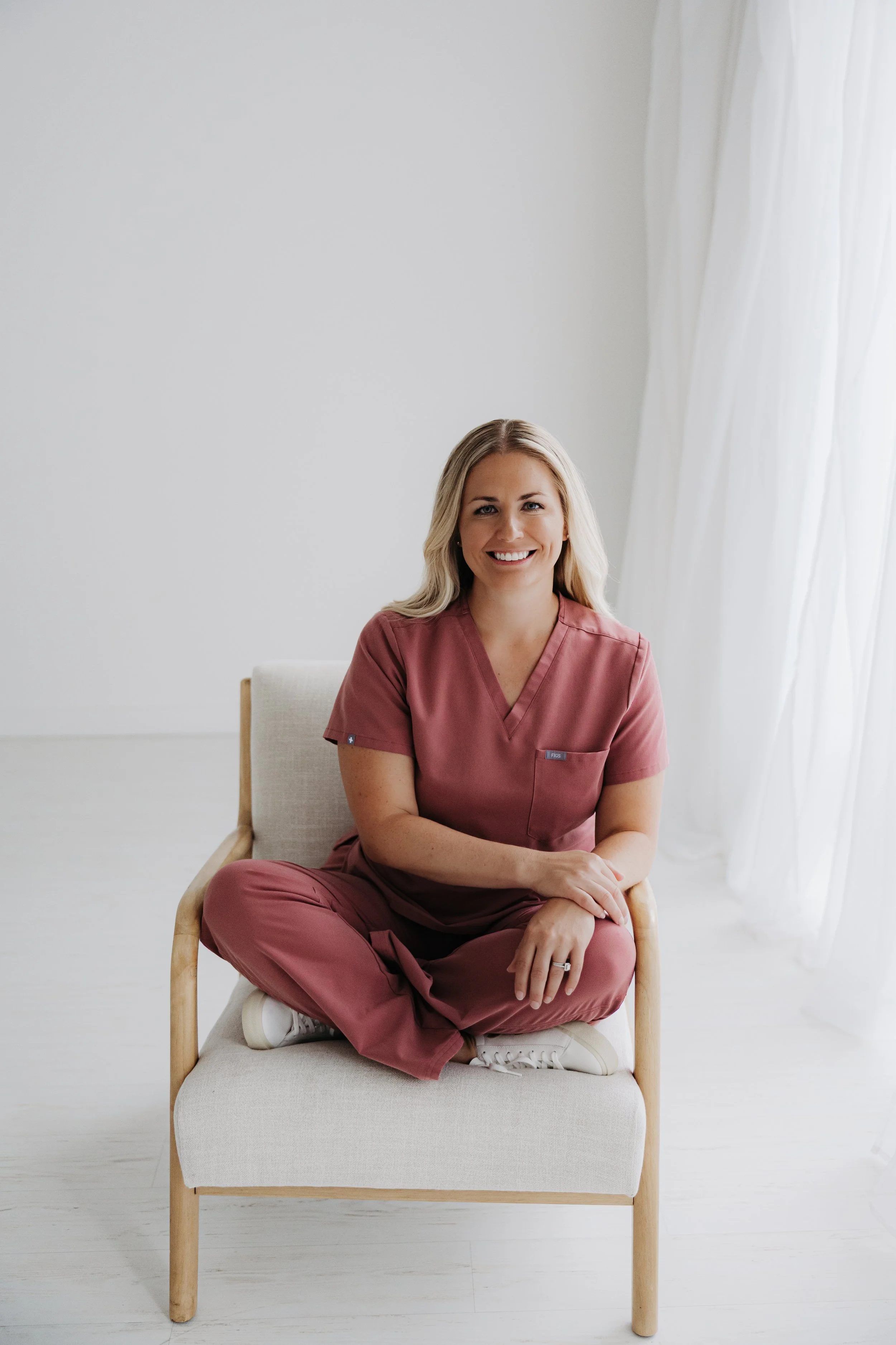 A smiling woman with long blonde hair, wearing pink scrubs, sitting cross-legged on an armchair in a bright, minimalistic room with white walls and curtains.