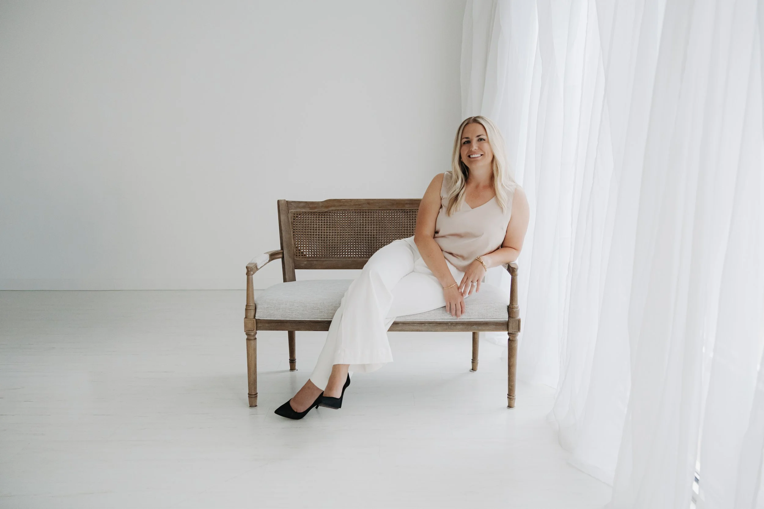 A woman with long blonde hair, wearing a sleeveless beige top and white pants, sitting on a wooden bench with a caned backrest. She is smiling, with her left arm resting on her lap and her right arm on the bench. She is wearing black high heels, positioned with her feet crossed at the ankles. The background is a minimalistic white wall with a sheer white curtain on the right side, allowing natural light to fill the space.