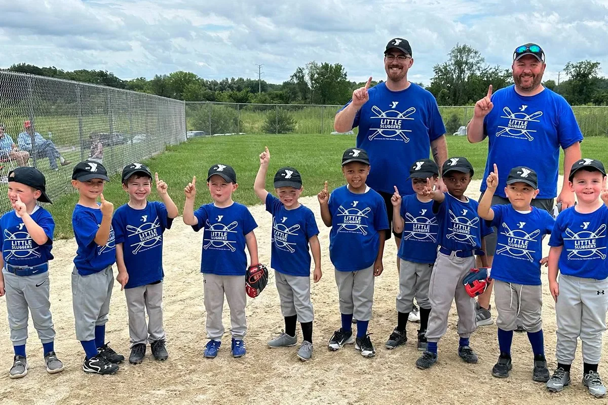 A group of young boys and their coaches at a baseball field, all wearing matching blue T-shirts and black caps, posing for a photo with their index fingers pointed up.