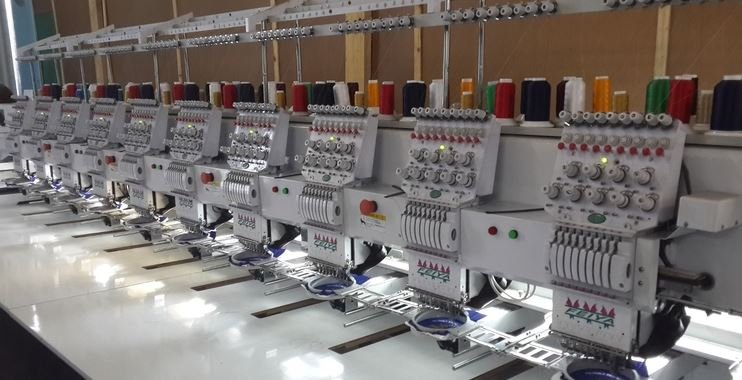 A row of industrial embroidery machines with thread spools on top, situated in a factory or workshop setting.