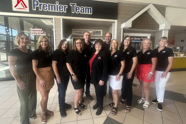 Group of nine women standing together inside a shopping mall in front of a sign that reads 'Premier Team,' some wearing black polo shirts, with two men in the group, all smiling.
