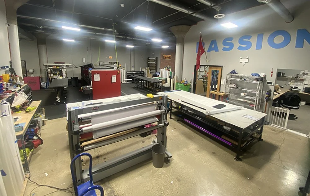 A workshop or manufacturing space with various workstations, cutting machines, tools, and shelves. The wall has large blue letters spelling 'PASSION'.