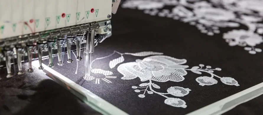 Close-up of an embroidery machine stitching a white floral design onto black fabric.