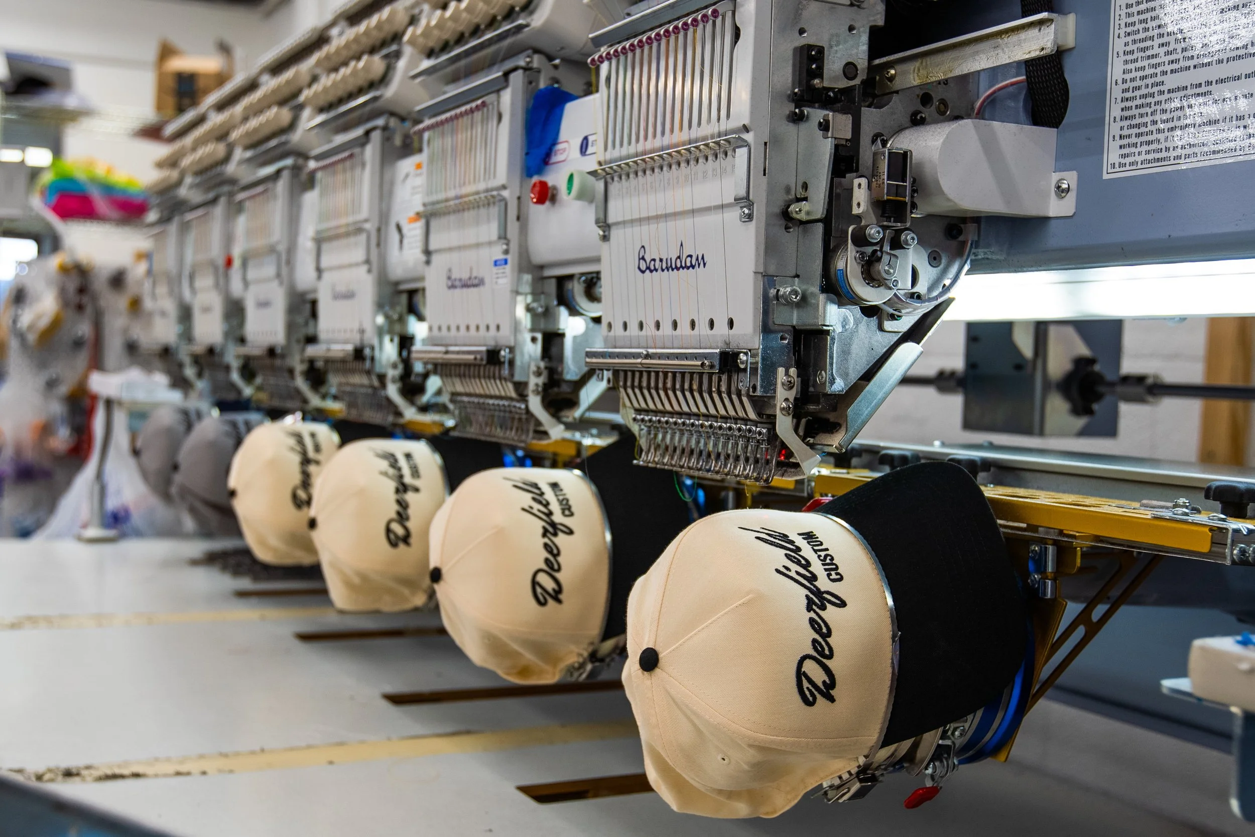 Close-up of an embroidery machine stitching the word 'Barudan' onto beige baseball caps, with multiple caps lined up and colorful threads in the background.