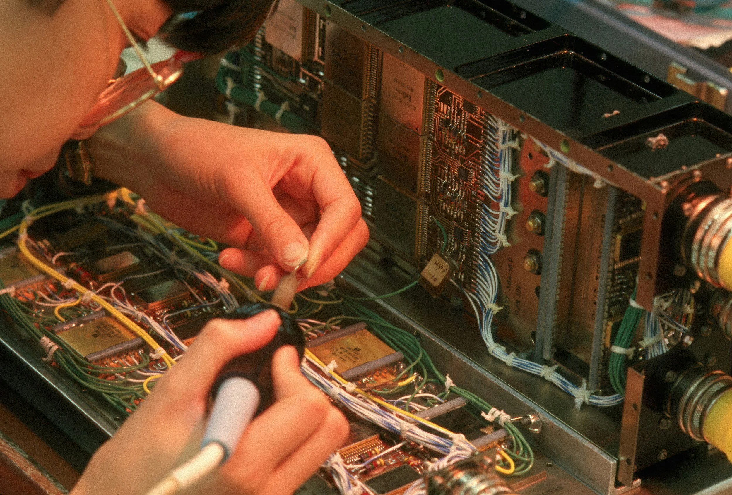 A person wearing glasses working on the interior electronics of a complex electronic device with circuit boards, wires, and components.