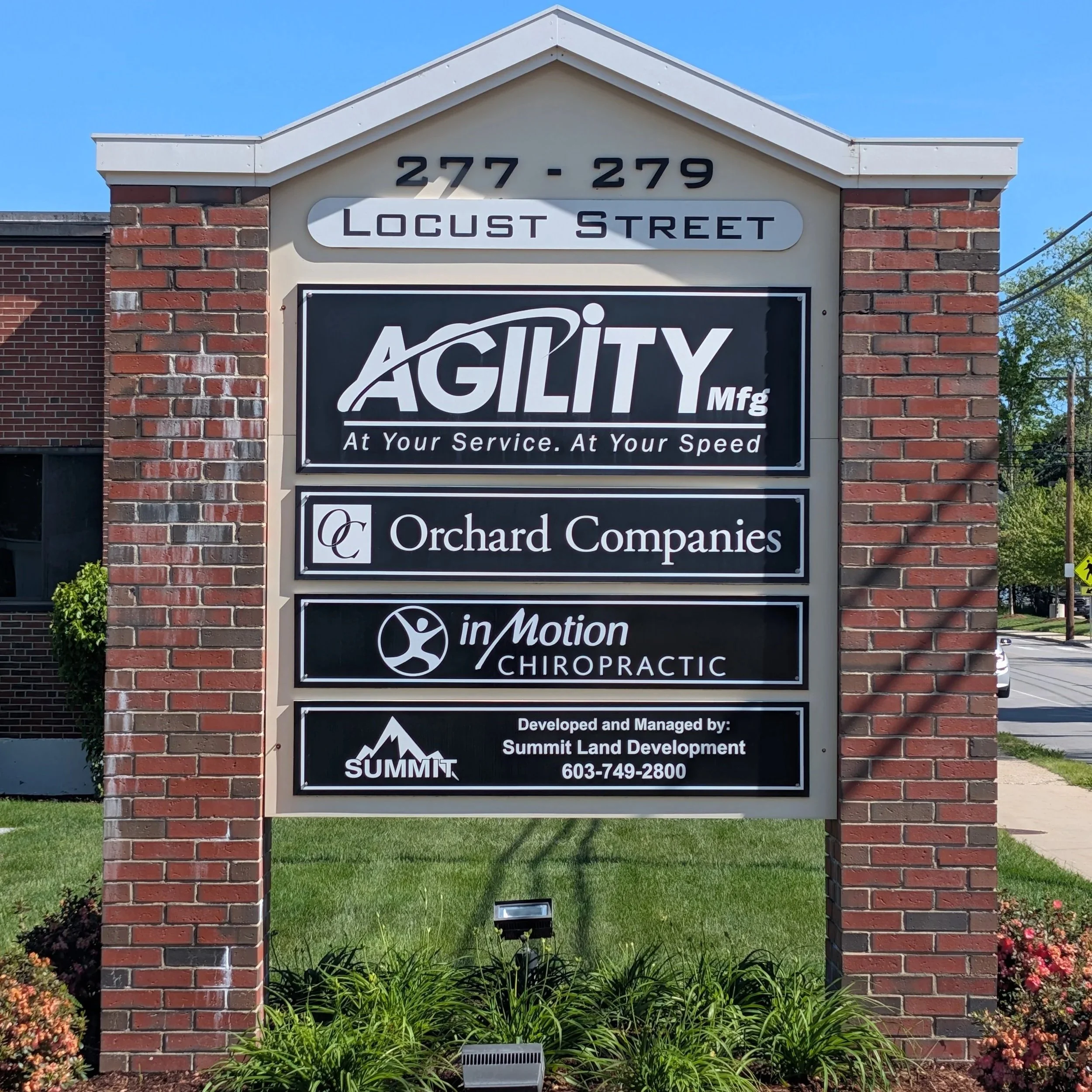 A framed business sign on brick pillars advertising companies located at 277-279 Locust Street, including Agility Manufacturing, Orchard Companies, and in Motion Chiropractic, with a well-maintained garden and green grass in front, under a clear blue sky.