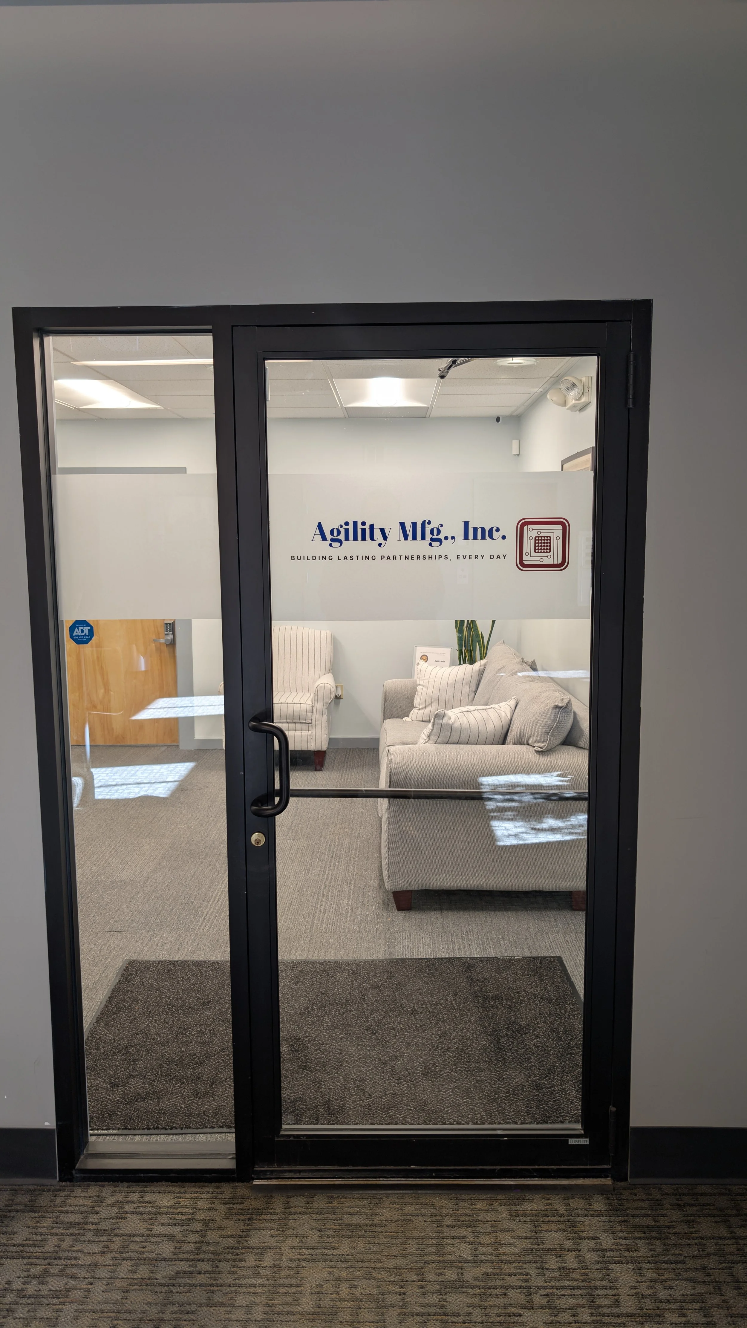 Office entrance door with a glass panel, displaying couches and a plant inside. The company's name on the door reads 'Agility Mfg., Inc.' with the tagline 'Building Lasting Partnerships, Every Day' and a logo of a calculator or device.