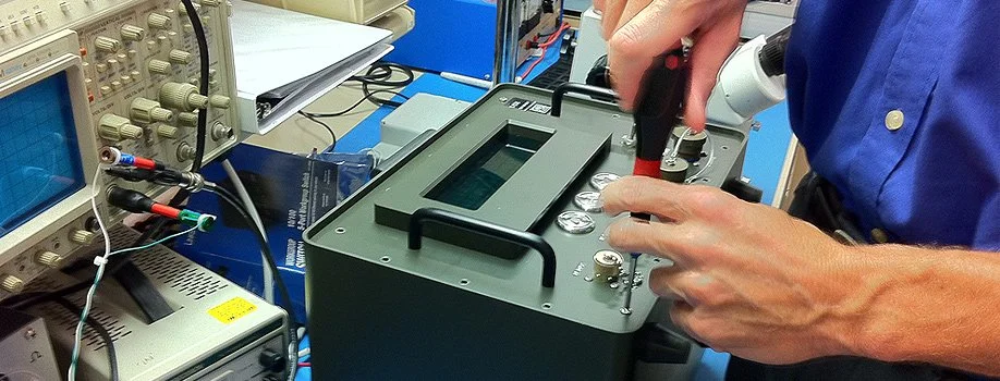 Person adjusting a piece of electronic testing equipment using a screwdriver in a laboratory setting with oscilloscopes and testing devices around.