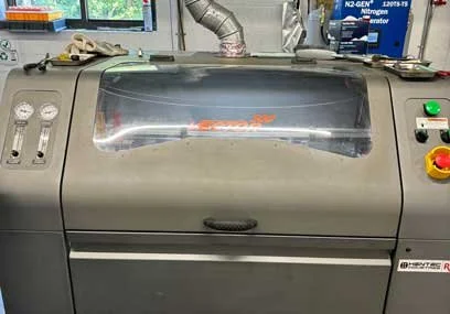 Industrial machine with control gauges and buttons, enclosed in a transparent cover in a workshop setting.