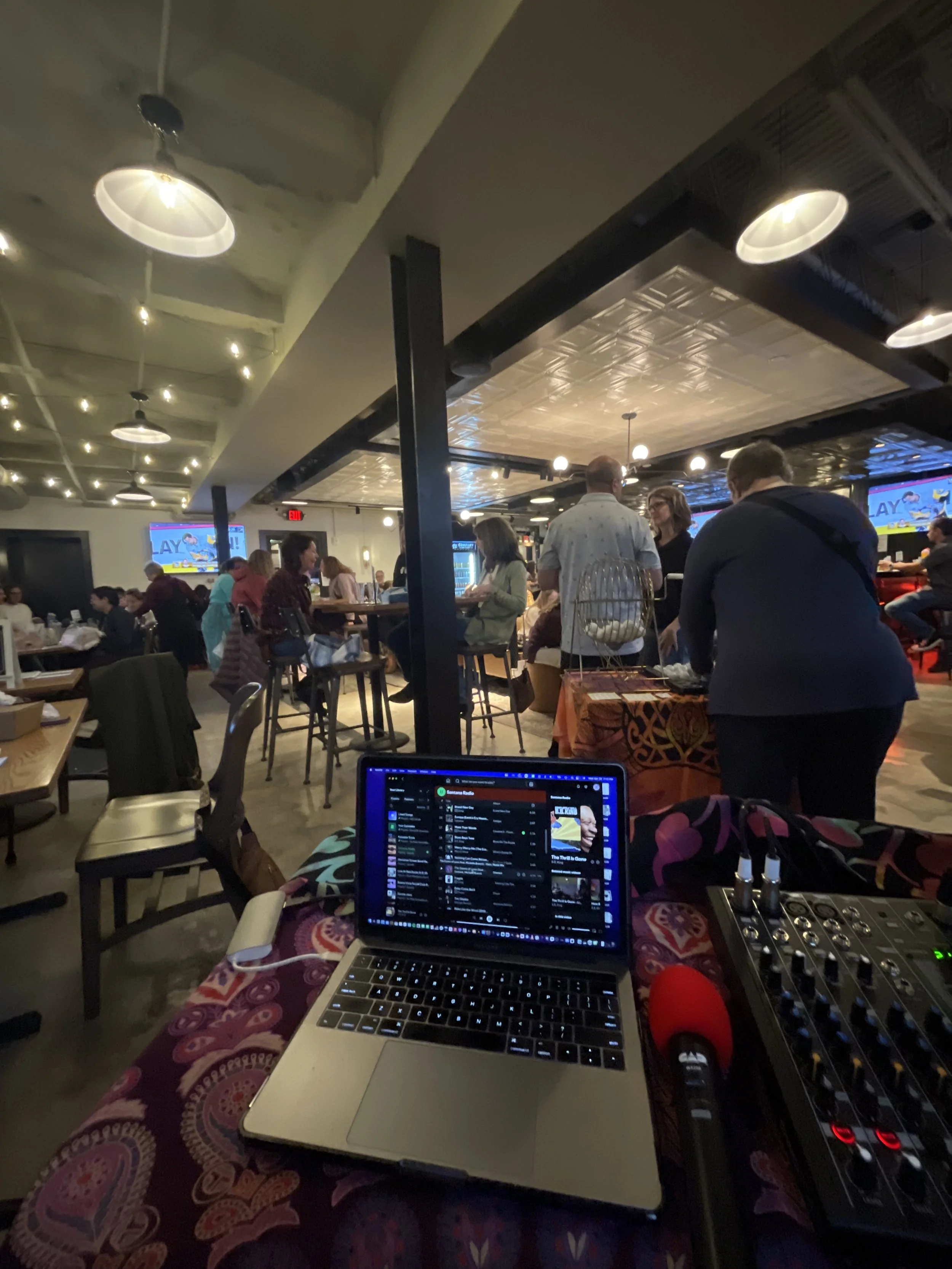 Indoor event or gathering with tables, people, and a DJ setup. Screen displays colorful graphics, and a laptop, audio mixer, and microphone are visible on a table in the foreground.